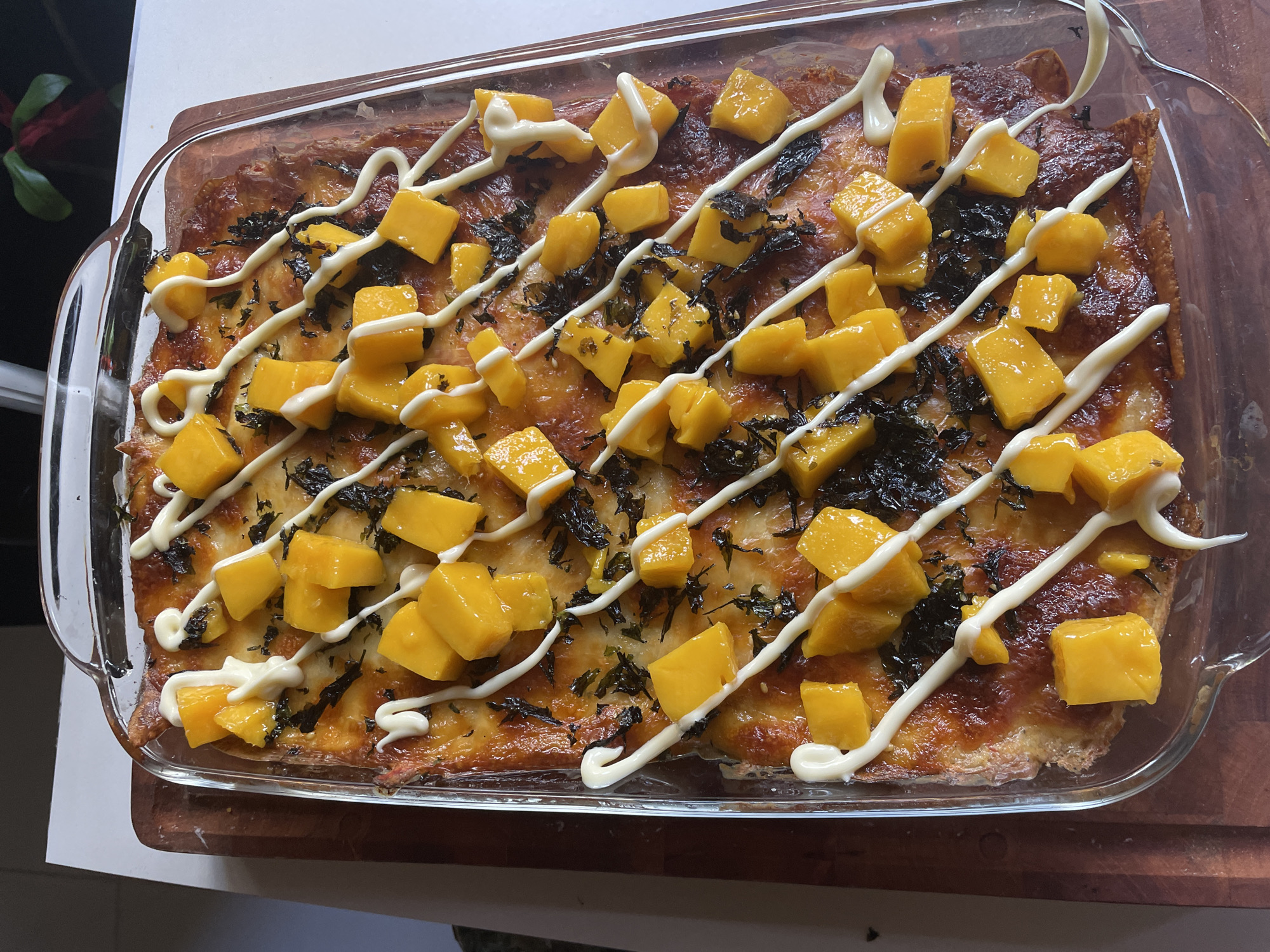 A glass baking dish sits on a wooden surface, containing a baked dish topped with diced mango, thin strands of what appears to be seaweed, and drizzled with a white sauce, possibly mayonnaise. The location is not discernible from the image.