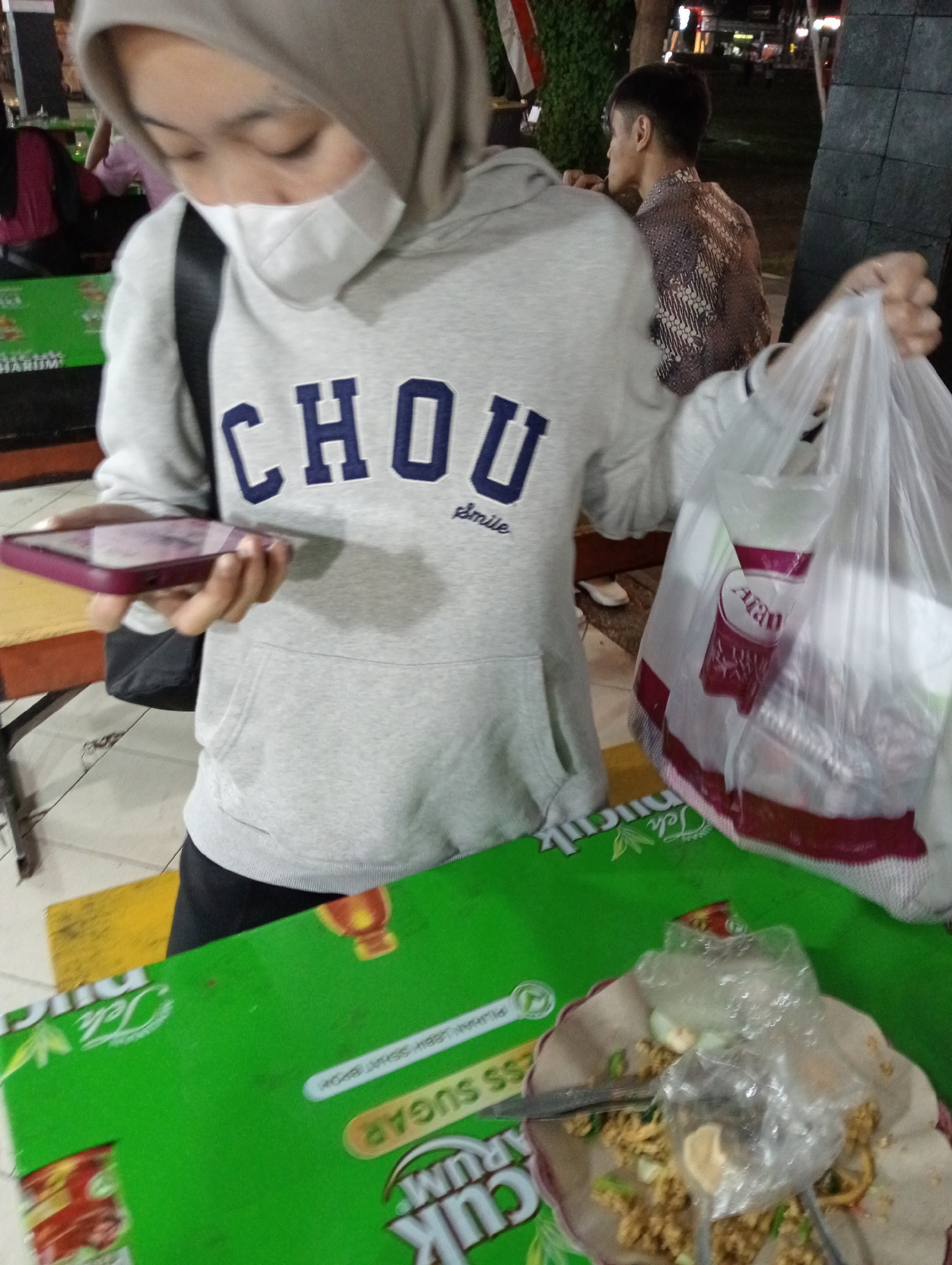 The image shows a close-up of a person wearing a light grey hoodie and a white face mask, looking down at a smartphone held in their left hand. The hoodie has the word "CHOU" printed in large, dark blue letters with a smaller word "smile" below it. The person is also carrying a transparent plastic bag filled with items in their right hand. In the background, another person wearing a batik shirt is visible. The scene appears to be outdoors at night, with artificial lighting. The foreground features a green table with some text and logos printed on it, as well as a plate with leftover food covered by a clear plastic wrapper. The environment suggests a casual dining or street food setting, possibly in Magelang, Indonesia, given the typical Indonesian food presentation and the presence of batik.