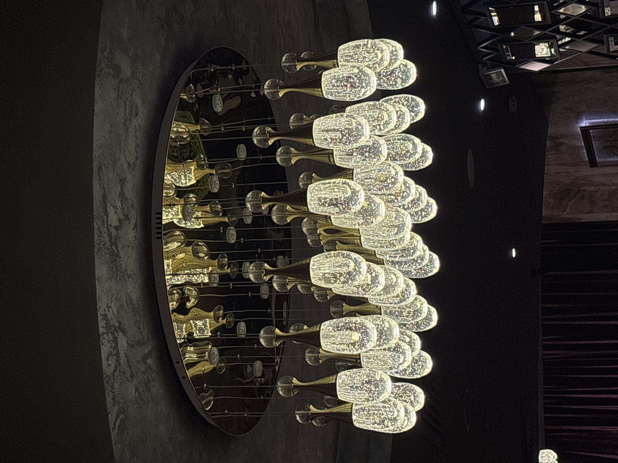 A view of a decorative circular chandelier mounted on a wall in a room. The chandelier consists of rows of illuminated glass fixtures, resembling champagne glasses, and is arranged in a reflective circular frame. The wall behind the chandelier has a mottled dark gray texture. In the background, a grid-like ceiling structure with recessed lights is visible, as well as a section of the wall with a striped pattern. The photograph appears to have been taken indoors, focusing on the lighting fixture as a key element of interior design. The location is Minsk, Belarus.