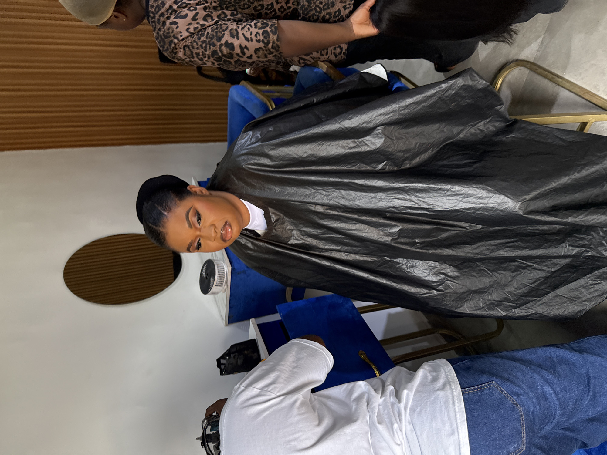 The image depicts a woman in a salon chair covered with a black styling cape. She has her hair styled and makeup done. A stylist wearing a leopard print top is holding a bundle of dark hair, possibly a wig or extension, behind her. Another person, wearing a white shirt and blue jeans, is standing near the chair, their upper body partially visible. The salon setting includes a small round window and a blue velvet chair. The location is likely a hair salon in Abuja, Nigeria.