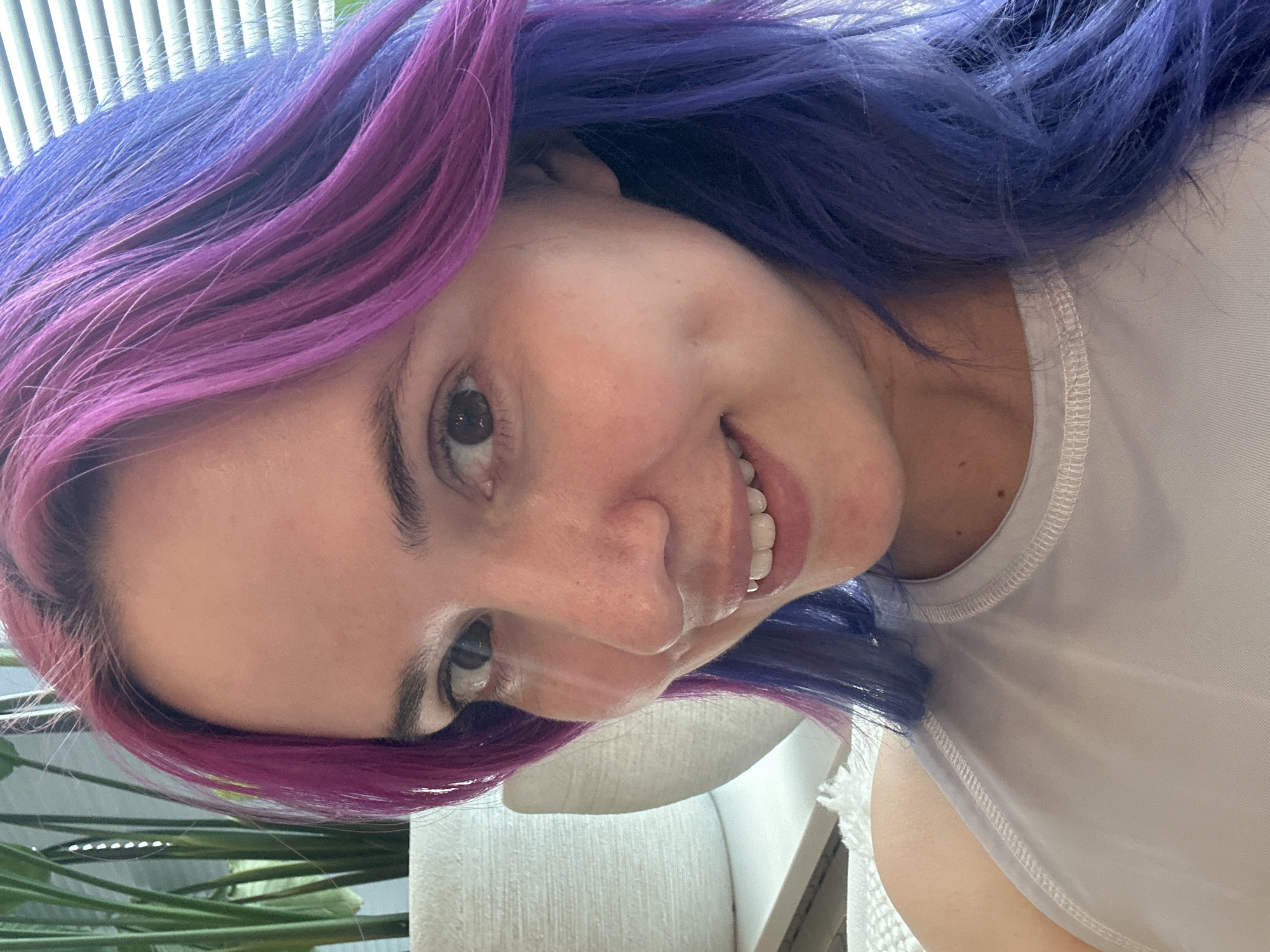 A close-up selfie shows a woman with vibrant, multi-toned hair, ranging from shades of purple to blue. She is smiling slightly. The background includes a window with blinds, some greenery (possibly a houseplant), and a partially visible white textured surface. The location is not discernible from the image.