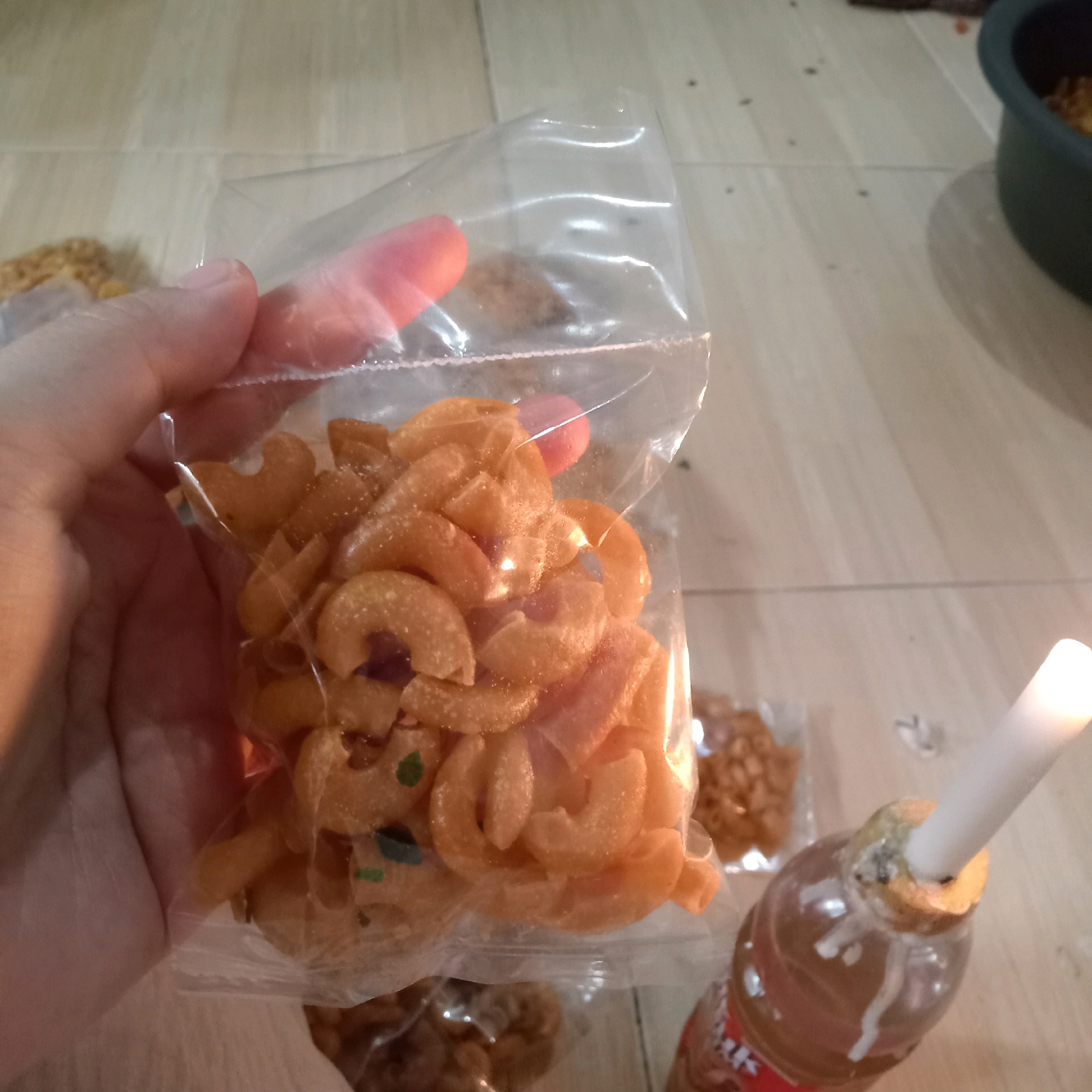 A close-up, eye-level shot captures a hand holding a clear plastic bag filled with orange, crescent-shaped snacks. The snacks appear to be fried and have a slightly powdery coating, with some small, dark purple and green flecks visible. The background shows a light-colored, tiled floor. To the right, a clear plastic bottle with a brown liquid and a white candle inserted into its opening is partially visible. The candle is lit, casting a bright glow. In the background, out of focus, are other small bags of snacks and a dark green bowl. The scene suggests an informal indoor setting, possibly a home, in Purwakarta, Indonesia.
