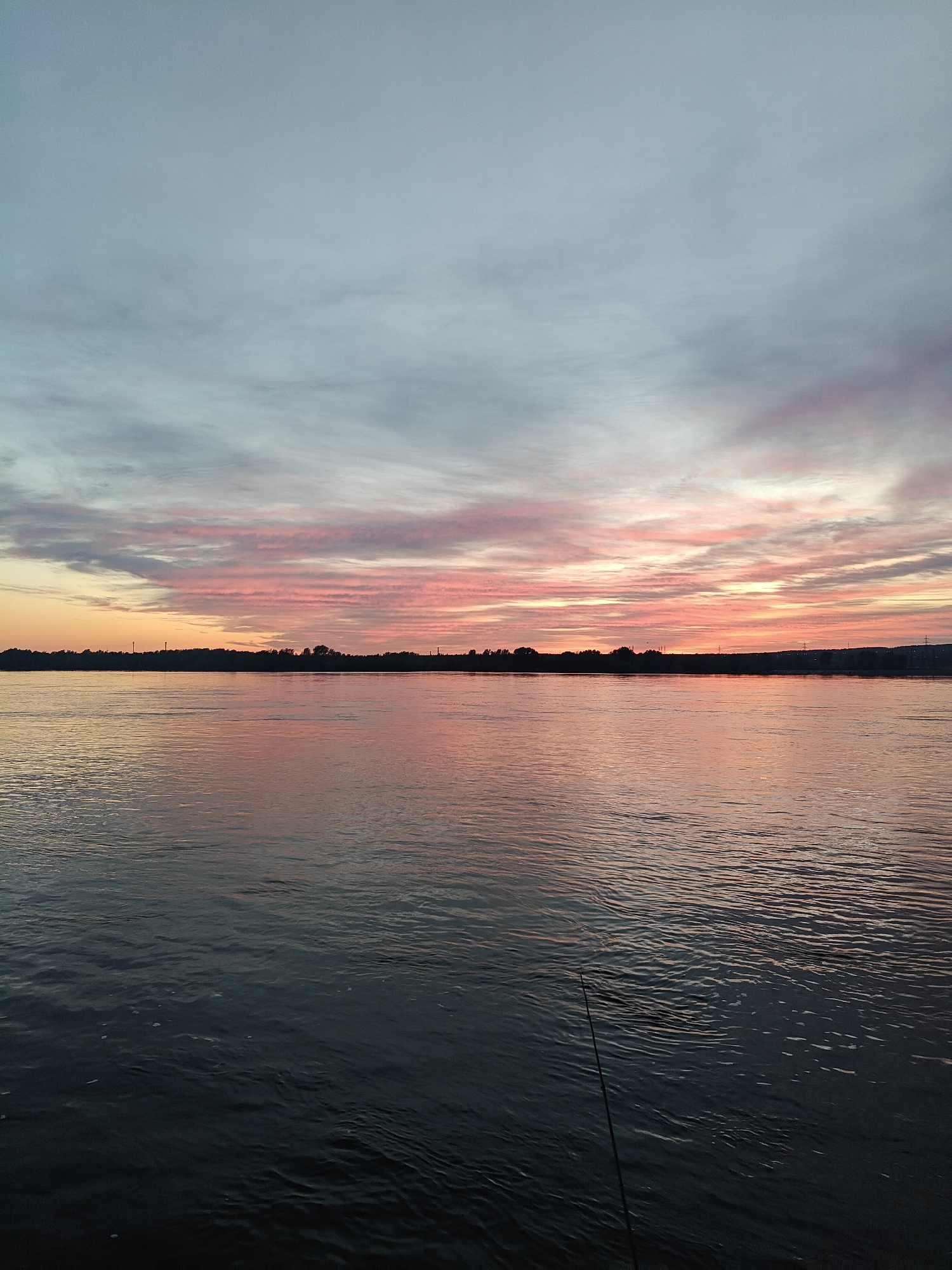 The image depicts a body of water, likely a river or large lake, under a dramatic sunset sky. The sky features a mix of pastel colors, including pink, orange, and blue, with cloud formations streaking across the horizon. The water reflects these colors, creating a shimmering effect. A dark, distant shoreline is visible, with what appears to be a faint outline of a city or settlement. In the foreground, a thin object, possibly a fishing rod, extends into the water. The location is likely Chaikovsky, Russia, based on the cityscape visible in the distance.