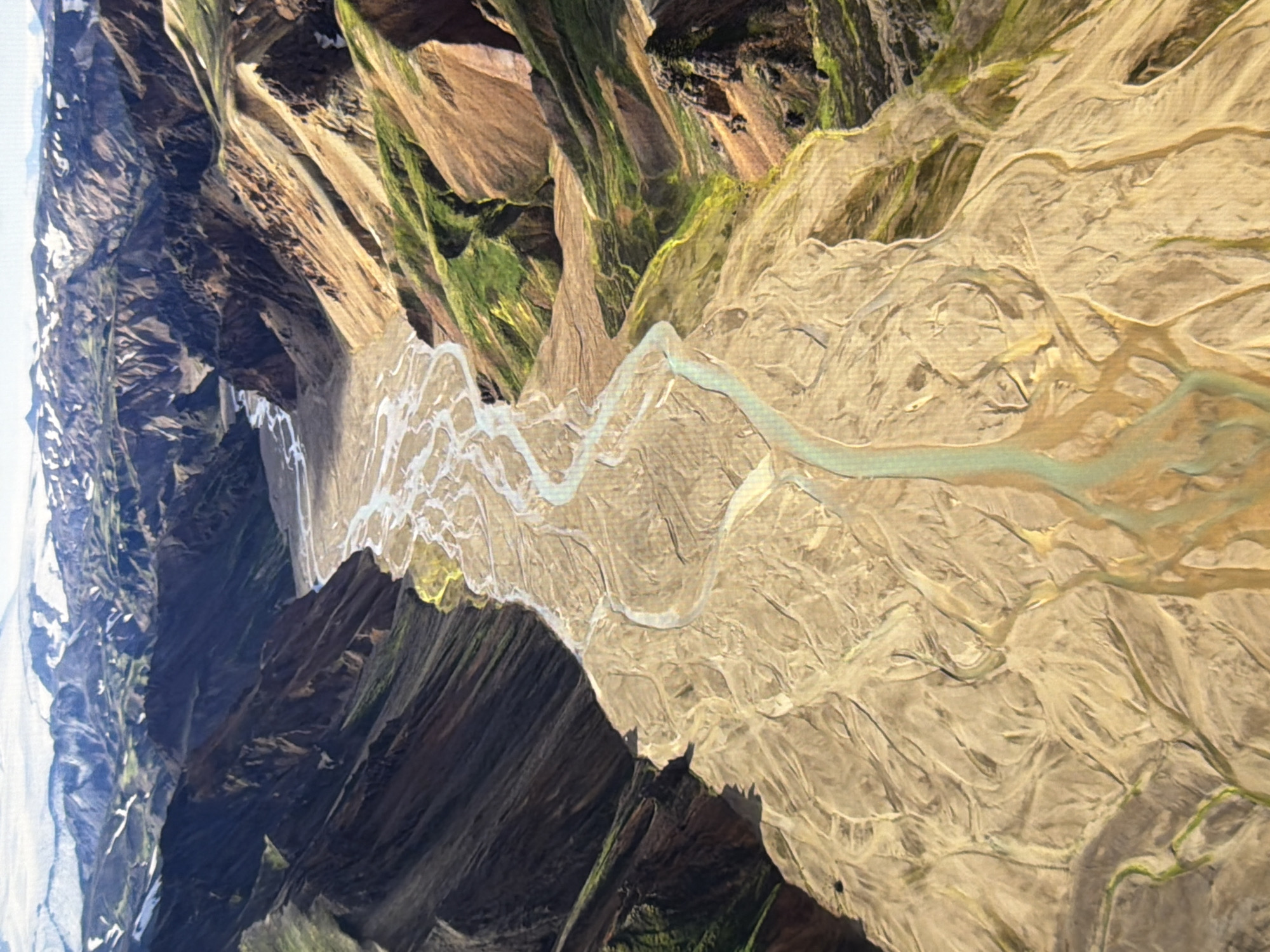 This is an aerial, wide-angle photograph of a dramatic, braided river valley surrounded by steep, colorful mountains. The river itself is wide and shallow, with a pale, sandy bed marked by numerous branching channels that split and merge. The surrounding slopes are a mosaic of earthy tones, ranging from ochre and brown to patches of vibrant green vegetation, particularly along the wetter gullies. In the distance, the mountain peaks are capped with snow and ice, under a hazy, bright sky. The overall impression is one of raw, natural power and geological time.
This image does not depict the city of Kyiv, Ukraine.