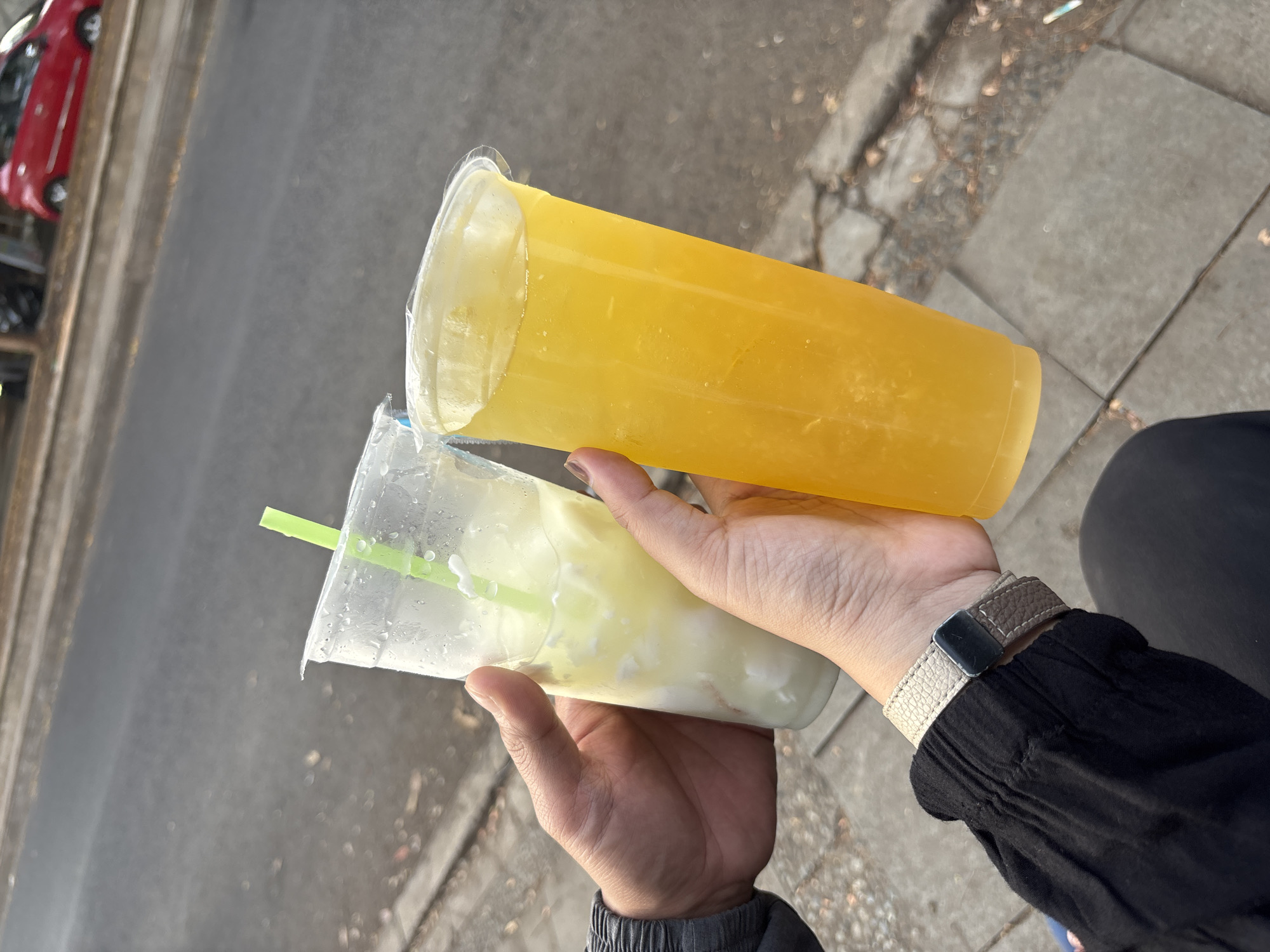 Two people are holding two plastic cups filled with beverages. The cup on the left contains a light-colored drink with solid pieces and a green straw. The cup on the right contains a bright yellow, translucent liquid. Both cups have clear plastic lids. The background shows a paved street with a red car parked on the side, indicating an urban setting in Cirebon, Indonesia. The ground is made of concrete slabs and asphalt. The image appears to be taken outdoors during daylight hours.