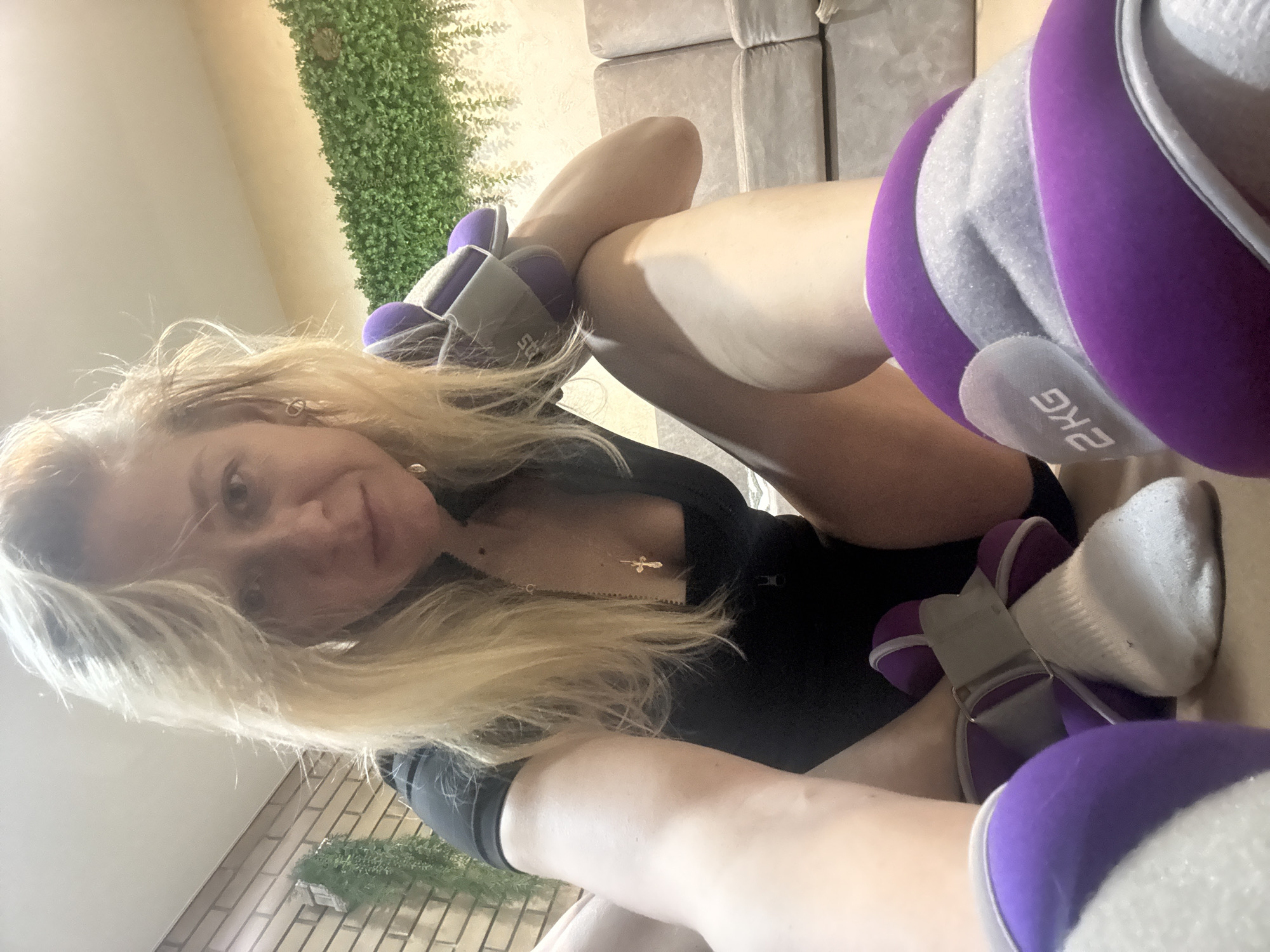 A woman is lying on a light-colored sofa, wearing 2 kg purple and gray weighted leg wraps. She is wearing a black top and has a cross necklace. Behind the sofa is a faux green wall. The location is unknown.