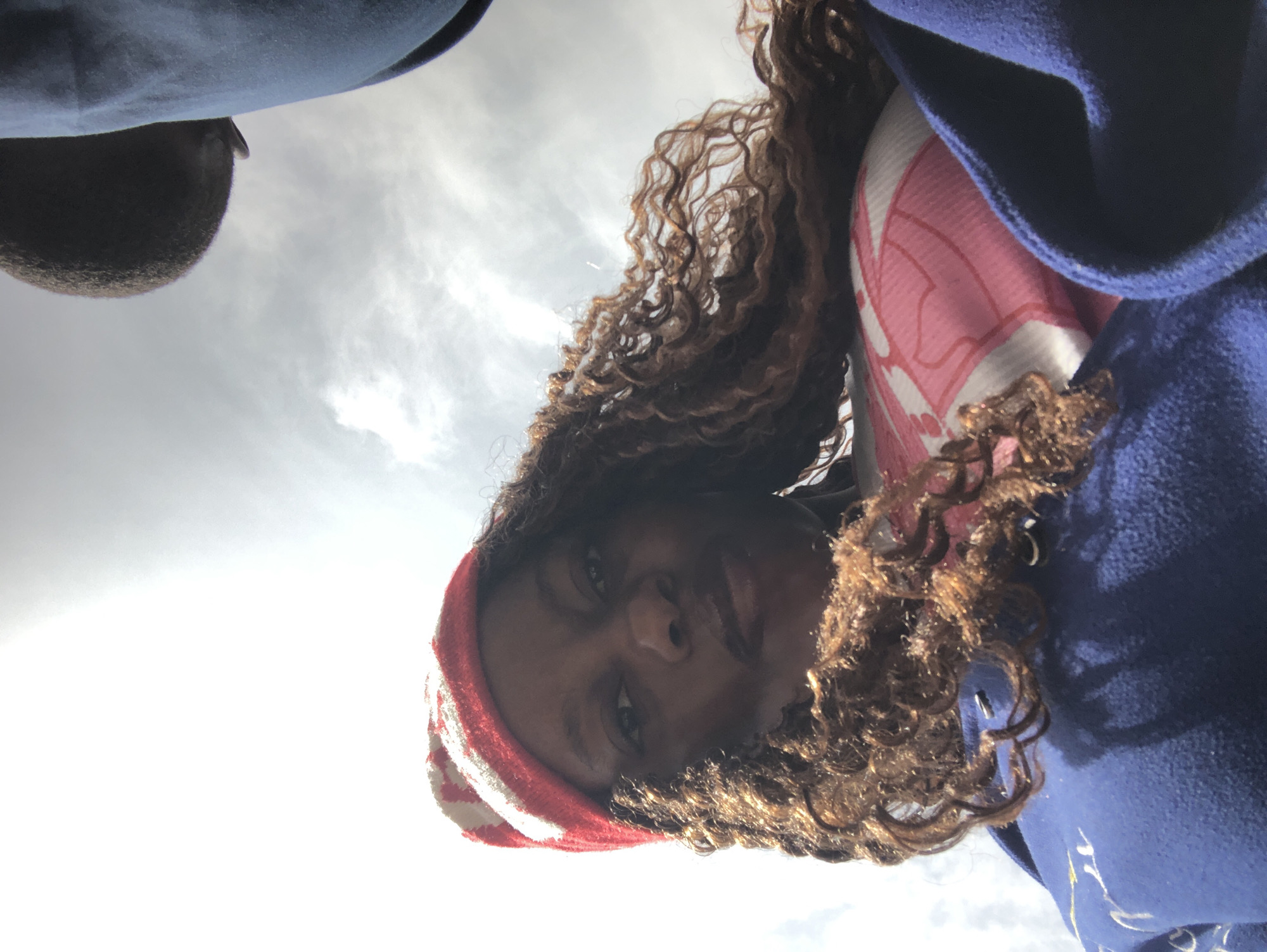 A low-angle shot looking up at a person with curly brown hair and dark skin, wearing a red and white striped headband and a blue coat. The person's face is visible, and they appear to be looking down at the camera. The background is a bright sky with some clouds. The location of the image is not discernible as Pindiga, Nigeria.