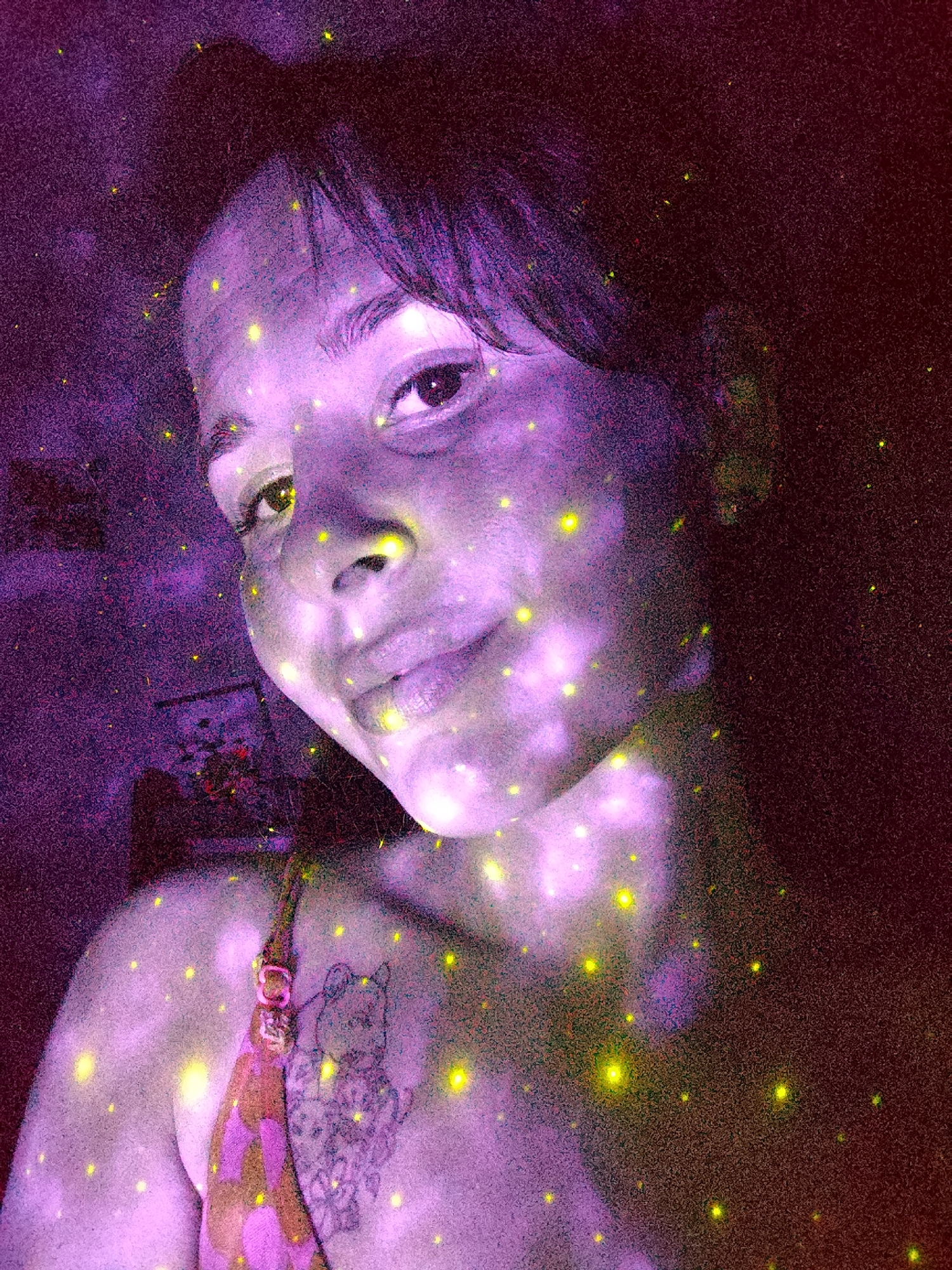 A person's face is prominently featured in the center of the image. The face is illuminated by numerous yellow speckles. A floral pattern is visible on the person's shoulder along with what appears to be a tattoo, also adorned with the same yellow speckles. The background is dark, with hints of a room interior and artwork visible. The overall color scheme leans heavily towards purple and red hues. It is not possible to identify the location as Фоки, Russia, with the information provided.