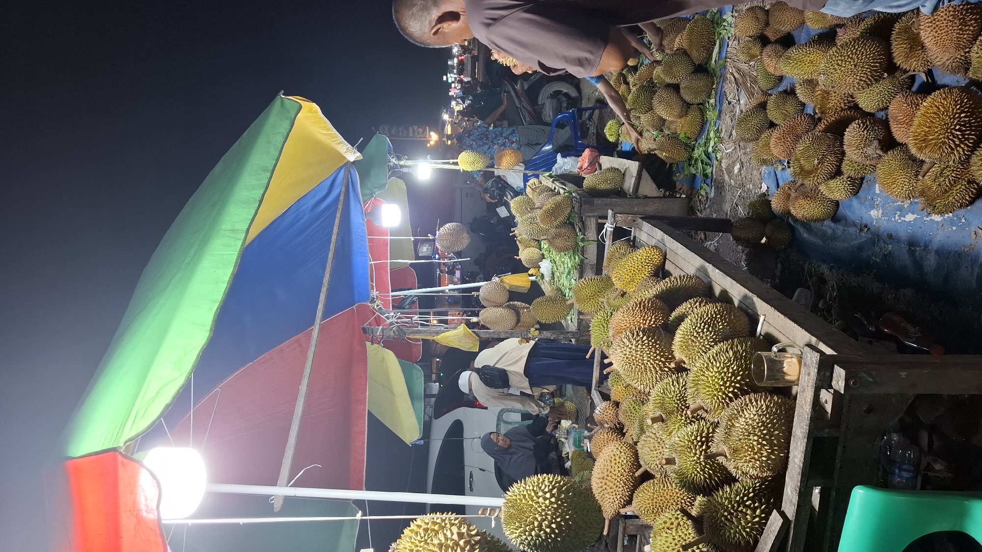 At night in Peurupok, Indonesia, a street vendor sells durian fruit under a colorful, multi-panel umbrella. The umbrella is supported by poles and lit by a bright overhead bulb. Piles of durian fruit are displayed on wooden tables and directly on the ground, some covered with blue tarps. Several people are present, including a vendor attending to customers and a man holding a durian. In the background, vehicles and motorcycles indicate this is a public street. The scene captures a lively market atmosphere.