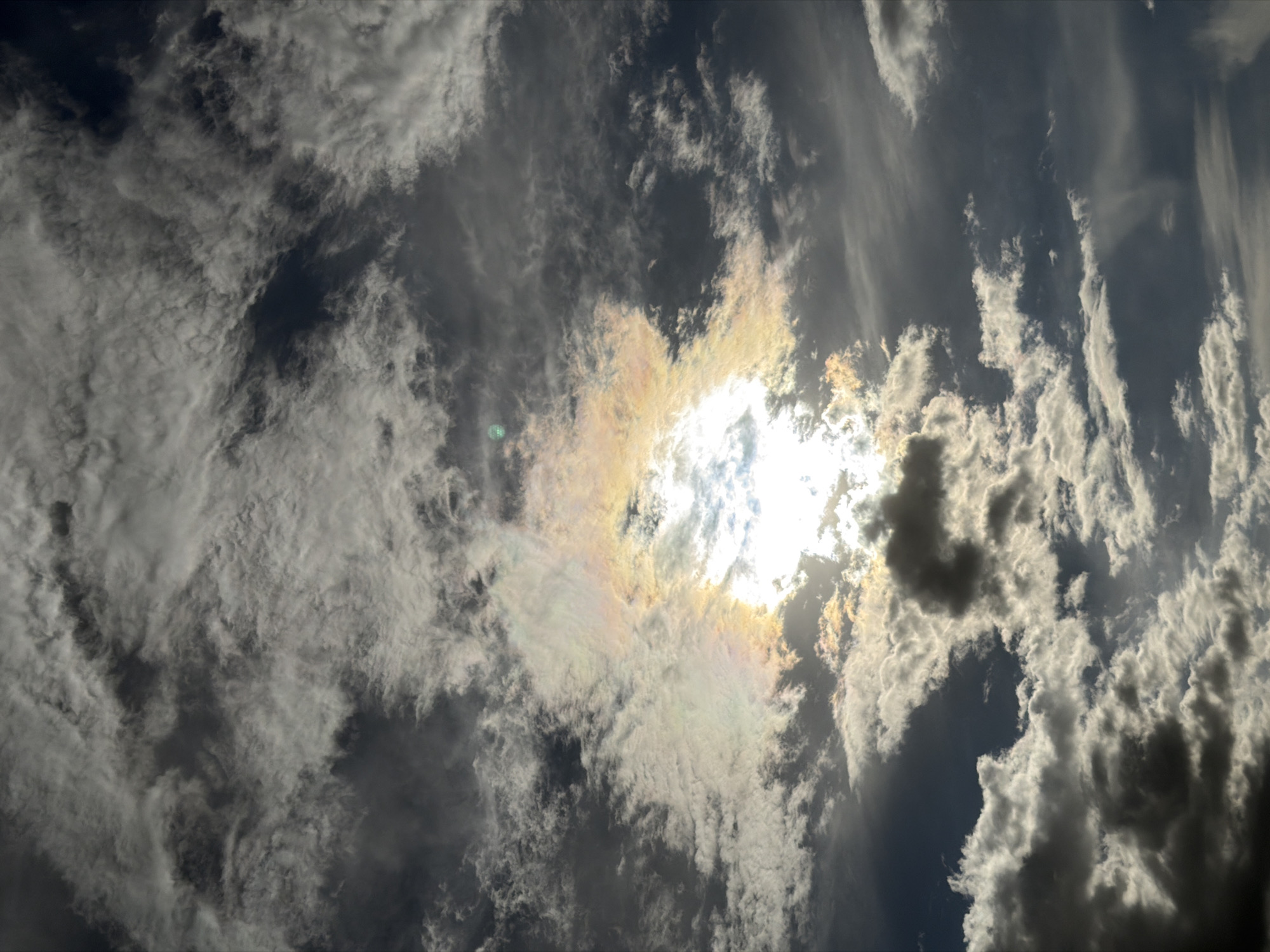 The image captures a view of the sky. A bright sun shines through a layer of clouds, creating a halo effect. The clouds are predominantly white and gray, with varying densities. The environment is the sky with clouds and sunlight. The location is not evident, as no city features are visible. The image depicts an atmosphere with weather-related subjects such as sunlight and clouds.