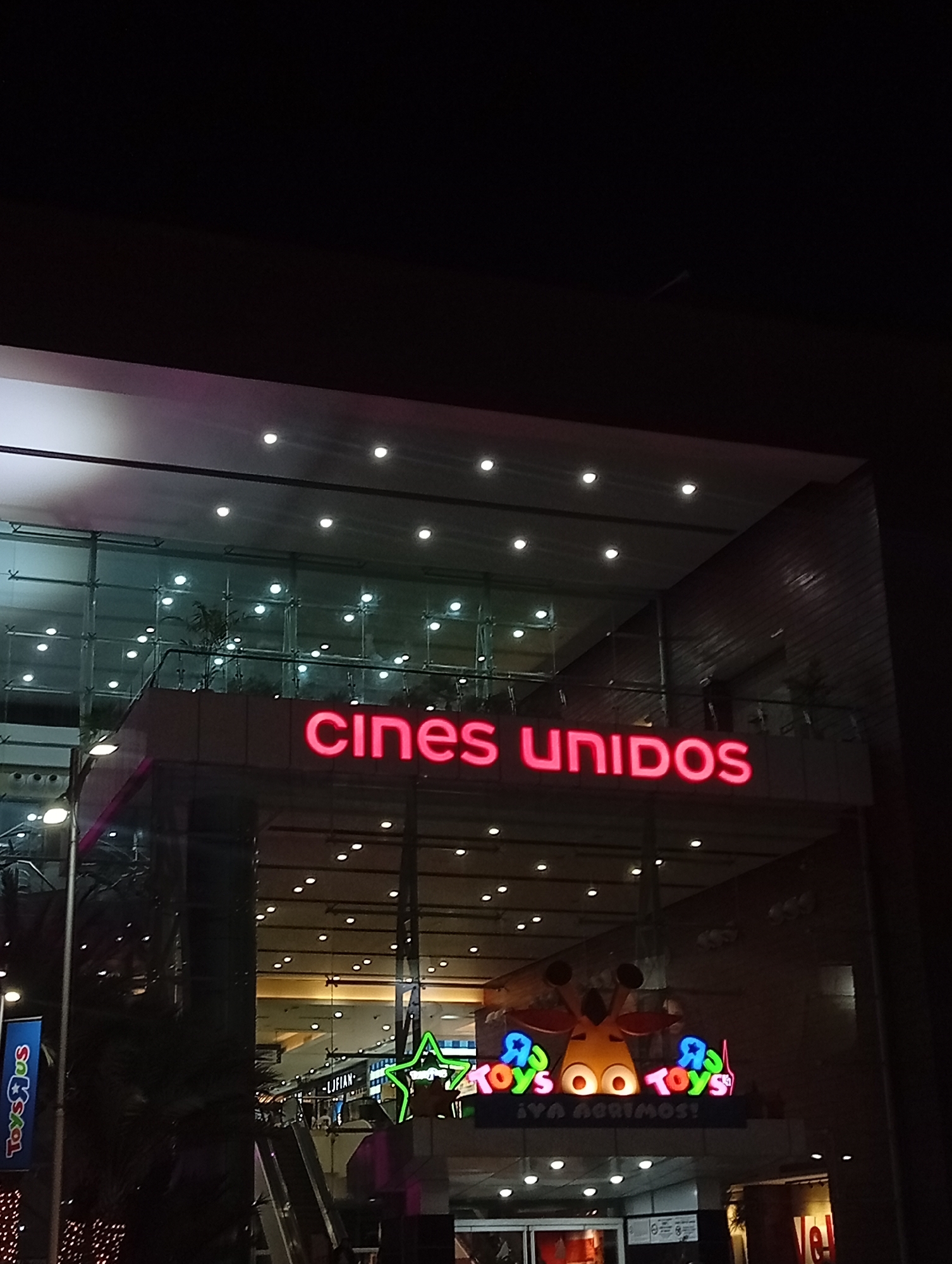 The image shows the exterior of a Cines Unidos movie theater located inside a shopping mall in Porlamar, Venezuela. The "Cines Unidos" sign is illuminated in red neon. Below the movie theater sign, a Toys"R"Us sign is visible. The mall's interior is visible through the glass facade, showing escalators, lighting, and storefronts. The image was taken at night.