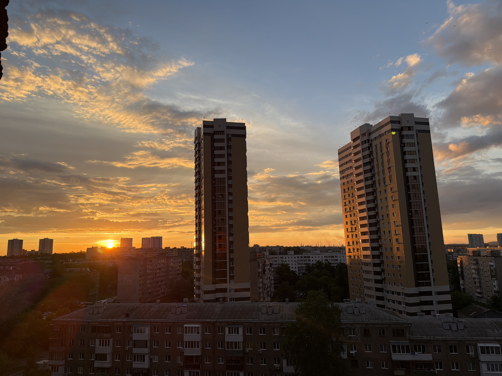 The image captures a vibrant urban sunset, illuminating a dense cityscape with warm, golden light. Two prominent high-rise residential buildings dominate the midground. The building on the left, slightly closer, features a patterned facade with light-colored horizontal bands and numerous windows, its reflective surfaces catching the intense orange glow of the sun. The second similar-style high-rise on the right also reflects the sunset, creating bright highlights on its windows and balconies.
The sun is visible as a bright, glowing orb positioned low on the horizon, towards the left side of the frame, peeking above distant buildings. Its powerful light bathes the sky in brilliant hues of orange, yellow, and soft pink, highlighting scattered clouds that streak across the heavens. Higher up, the sky transitions to a soft blue.
Below and between the prominent high-rises, a sprawling urban environment unfolds, composed of numerous lower-rise apartment blocks, some with visible balconies and windows. Many of these buildings appear older, with darker tones, and are partially shrouded in the long shadows cast by the low sun. Patches of green foliage from trees are interspersed throughout the urban fabric, adding a touch of nature to the concrete landscape. The overall scene conveys a sense of peaceful twilight, with the dramatic colors of the setting sun creating a striking contrast against the architectural forms. No discernible text is visible in the image.