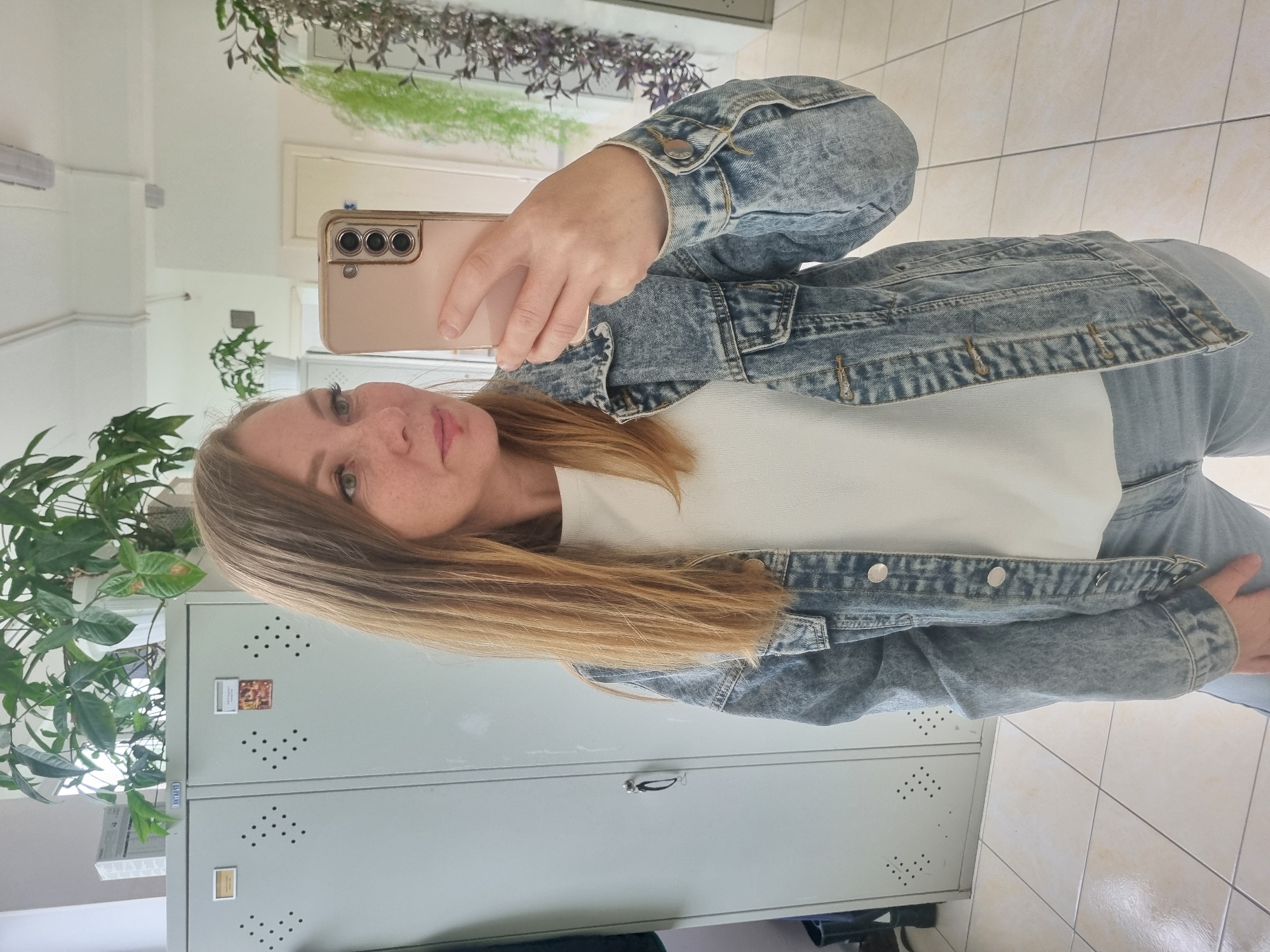 An indoor mirror selfie captures a woman holding a light-colored smartphone, possibly a Samsung Galaxy, to take her picture. She has long, light brown hair, light eyes, and freckles, with a neutral expression. She is wearing a light-wash denim jacket over a white t-shirt, paired with light-colored jeans.
The setting appears to be a well-lit interior space, potentially an office, school, or public building hallway in Kemul, Russia. Visible in the reflection behind her are several grey metal lockers. One locker displays a small square picture or card, and a smaller black and white sticker that appears to say "LIVELY LINK" in its bottom left corner. A small keyring or charm hangs from one of the locker handles. Green potted plants are strategically placed throughout the area, adding a natural element. The floor is covered with light-colored square tiles, and the walls are white. No specific time of day or weather is evident.