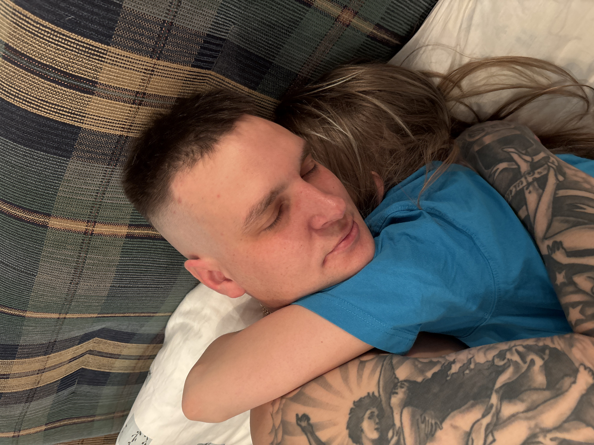 The image depicts two individuals, an adult male and an adult female, in a state of repose within an indoor setting in the city of Русский Вожой, Russia. The male, identifiable by his short-shaved haircut, closed eyes, and fair skin, wears a blue short-sleeved t-shirt and a thin metallic chain around his neck. His left arm, extensively covered with black and grey tattoos depicting figures and patterns, is wrapped around the female. The female, whose face is largely obscured by her long, light brown hair with subtle highlights, is nestled against the male's chest/shoulder. Her right arm, also tattooed with dark designs, is partially visible. Both individuals are lying on white bedding, set against a background of plaid fabric featuring dark green, brown, and blue stripes.