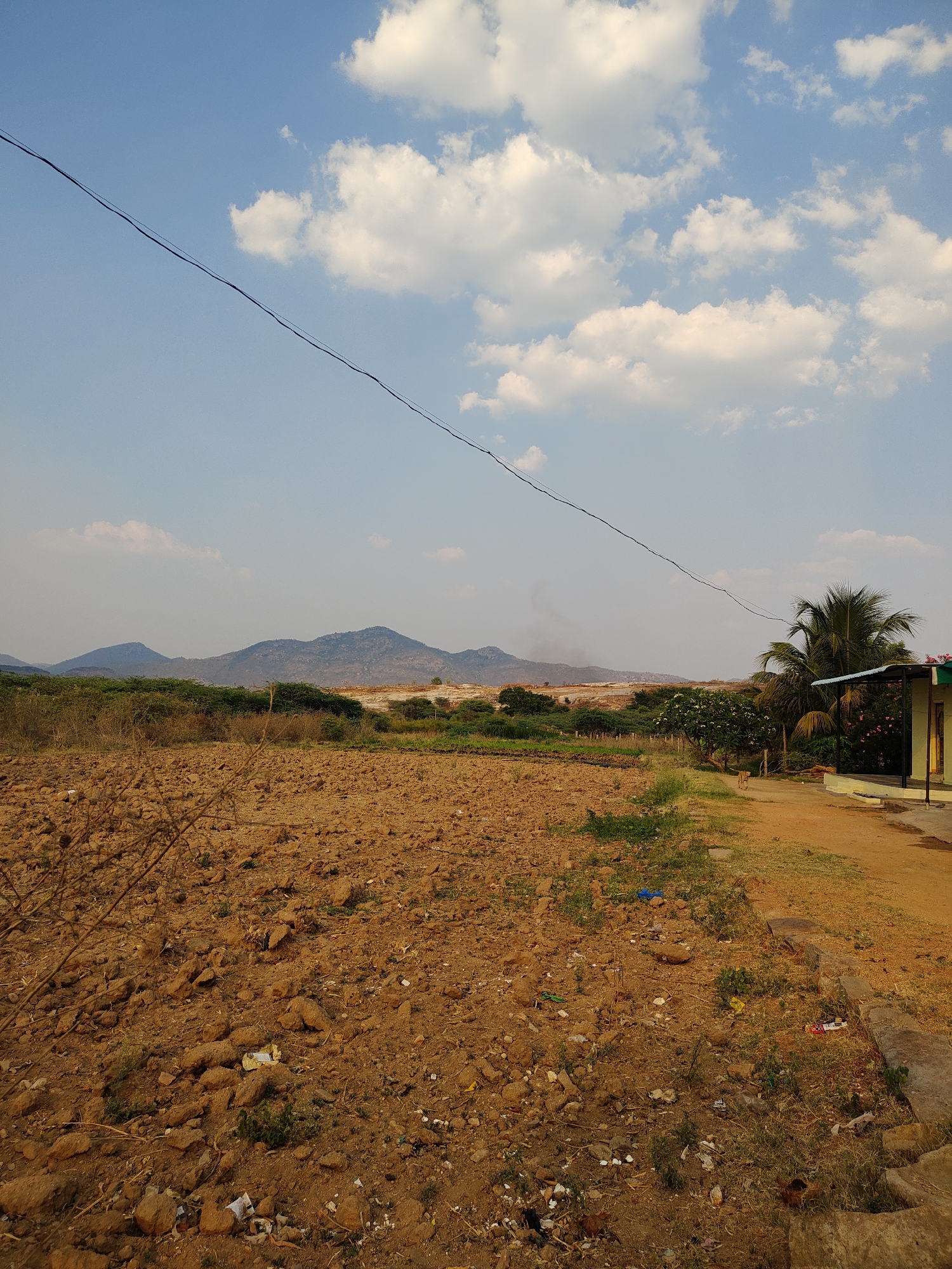 The image depicts a rural scene near Kammavaripalli, India. The foreground shows a field of dry, brown, rocky soil with sparse vegetation. In the middle ground, there is a strip of green vegetation, followed by a building with a green roof and a few trees, including a palm tree. In the background, there are hills and a large quarry. The sky is blue with scattered white clouds. A powerline stretches across the top of the image. No people are visible in the scene.