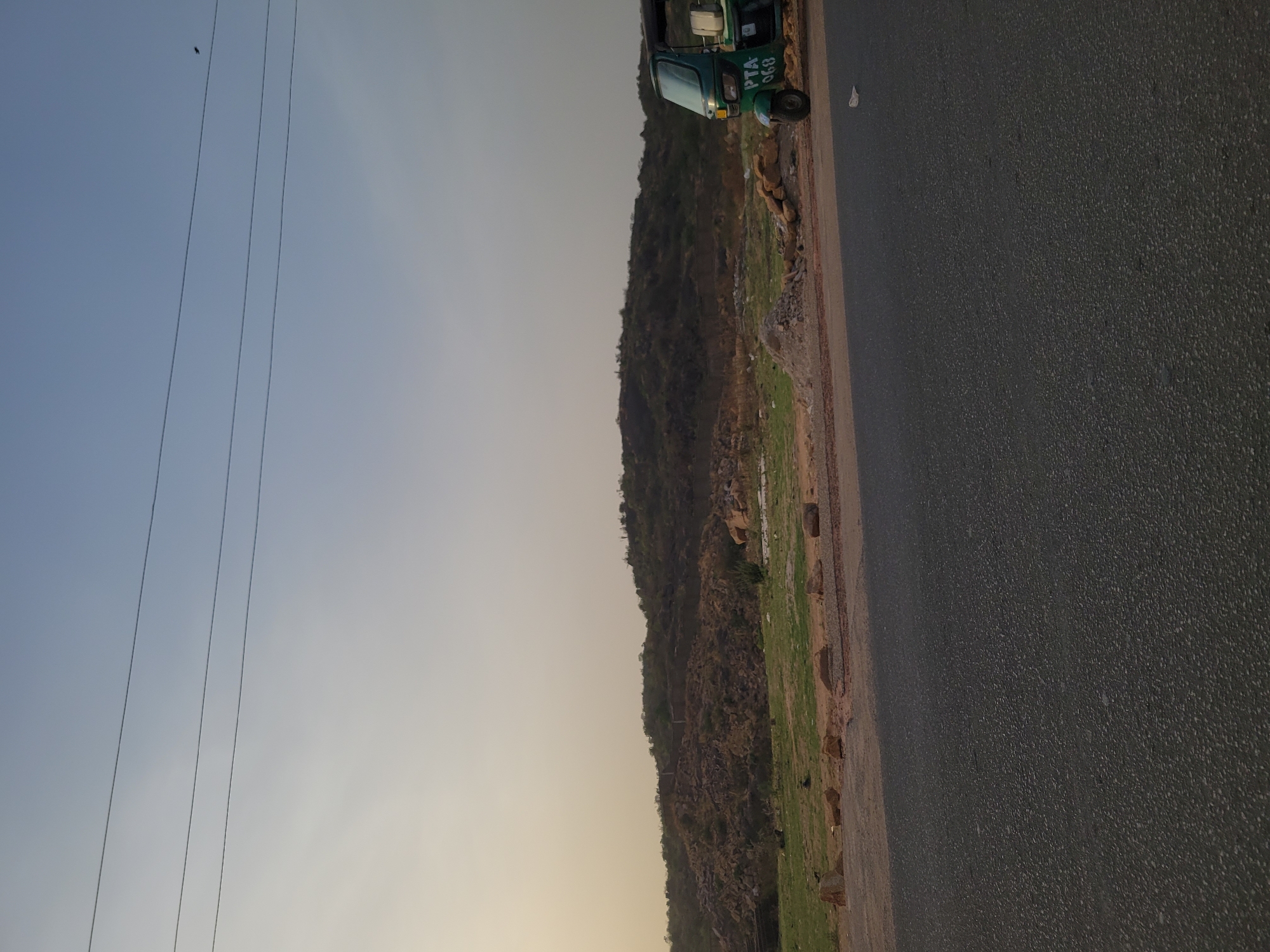 The scene is a roadside in Jos, Nigeria, showing a green three-wheeled vehicle (auto-rickshaw or "Keke Napep") parked near a road. There is a hillside with visible rock formations and sparse vegetation next to the road. The sky is visible above the hillside, with power lines crossing the frame. The road surface is asphalt.