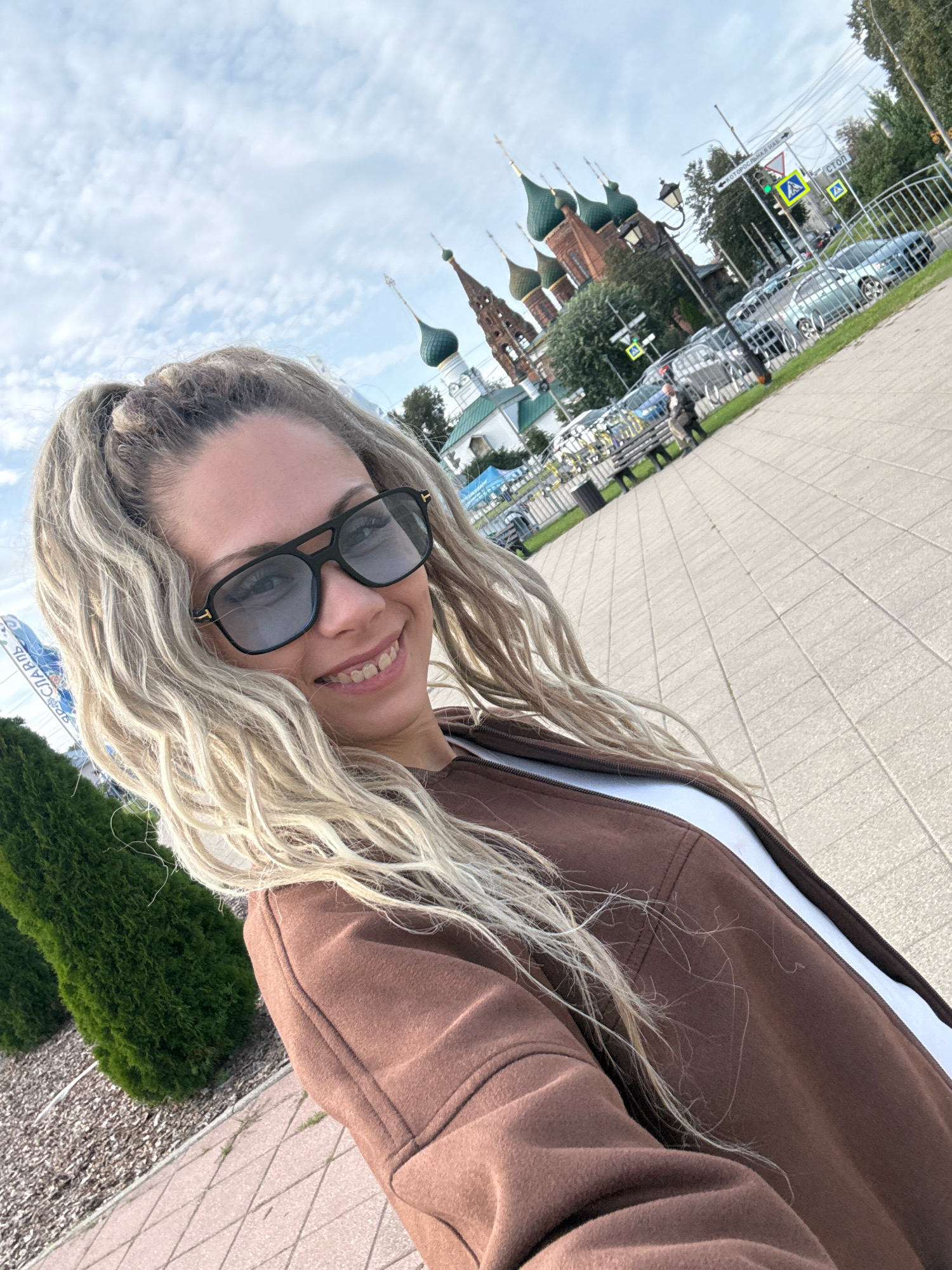 A woman with blonde hair pulled into a high ponytail and wearing black-rimmed sunglasses takes a selfie outdoors. She is wearing a brown jacket over a white top. Behind her, a church with green domes and a tall bell tower is visible. The church is surrounded by trees and a street with cars. The ground is paved with light-colored square tiles. The sky is partly cloudy.
