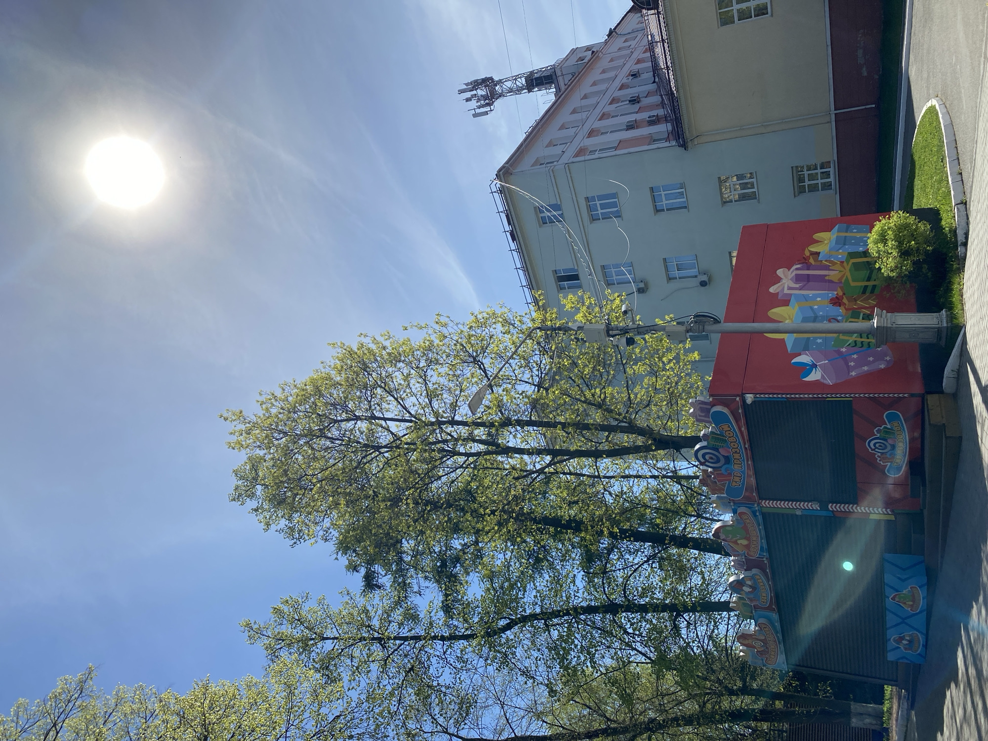 The image depicts a bright, sunny day in what appears to be a city. The sky is blue with the sun visible in the upper left corner. A tall building is present in the background, with a cellular tower mounted on its roof. A kiosk, brightly painted with gift box imagery on its upper section and food-related designs on the lower section, stands in the foreground. Trees with fresh green leaves partially obscure the building and the kiosk. The location is suspected to be Izhevsk, Russia. There are no people visible in the image.