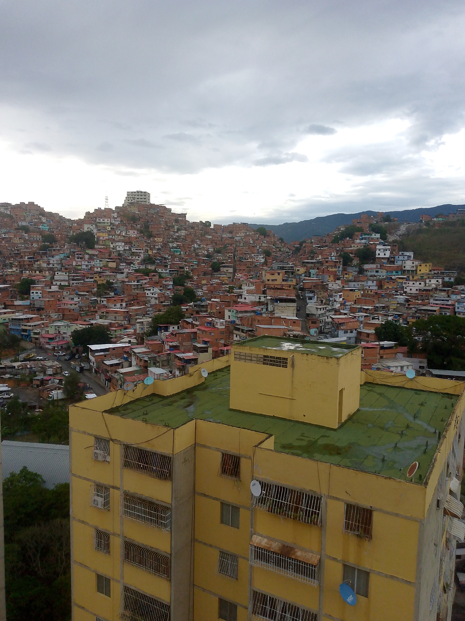 This high-angle, panoramic image depicts a densely populated urban landscape in Caracas, Venezuela, under an overcast sky. The foreground features the upper floors and rooftop of a multi-story, yellow-painted concrete building. This building has multiple windows, many secured with white metal grilles, and some visible air conditioning units. Its flat roof is coated in a weathered green material, showing dark moisture stains, and features small structures, utility lines, and several satellite dishes, including one white dish and one blue dish branded "inter."
Beyond the foreground building, the midground and background reveal extensive informal settlements, characteristic of Caracas's "barrios" or "ranchos," sprawling across numerous hillsides. These settlements consist of countless small, closely packed houses, predominantly constructed from unpainted red brick, with various colored facades (including blue, white, and yellow) and a mix of roof types, often corrugated metal. A taller white building is visible on a prominent distant hilltop, along with a communication tower. Vehicular roads and pathways are discernible winding through the dense housing clusters. The topography is distinctly mountainous, with additional tree-covered hills and a mountain range forming the distant horizon. No individual people are distinctly visible. The sky is predominantly gray and cloudy.