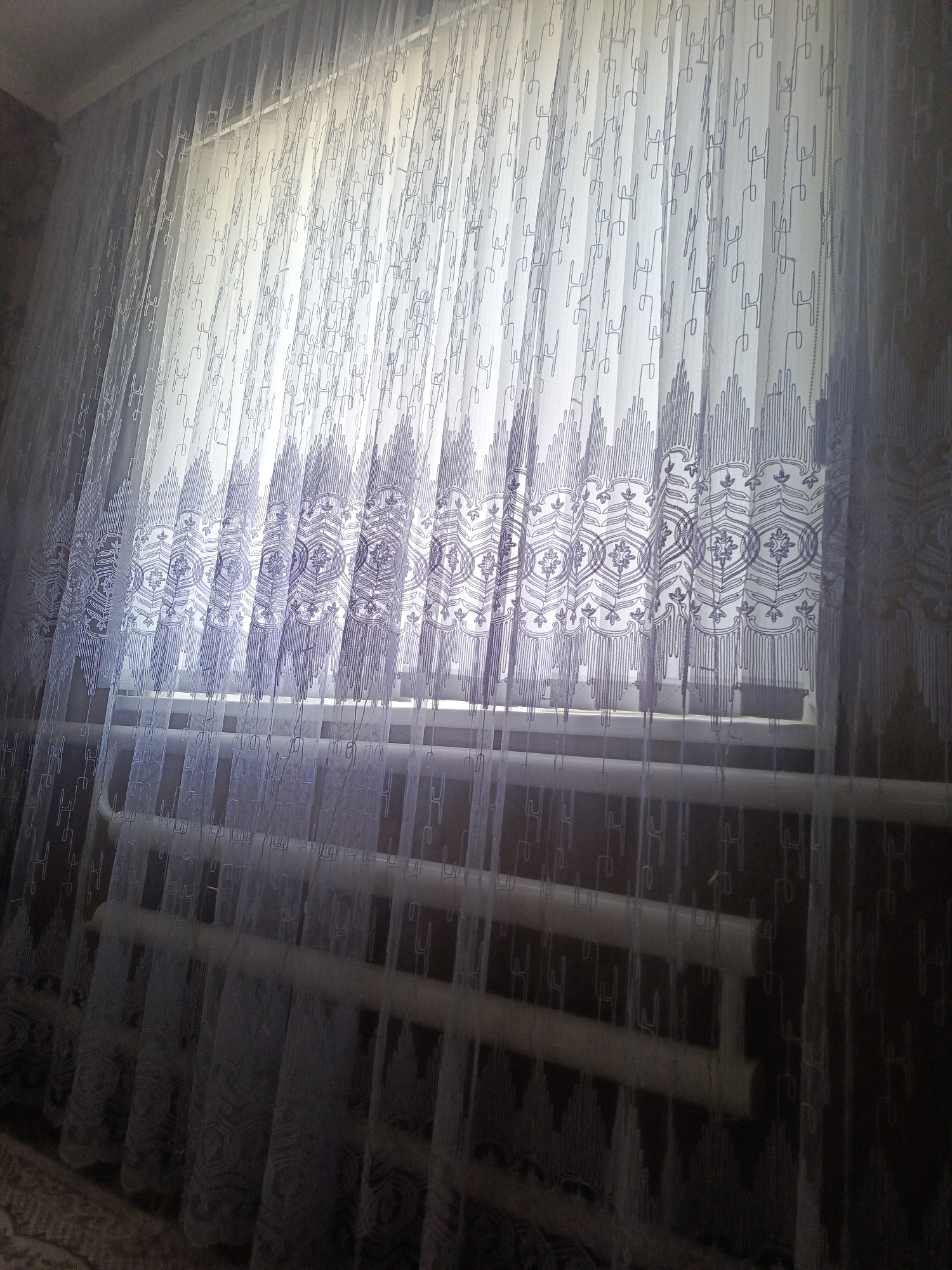 The image shows a close-up view of sheer curtains with an intricate patterned design. The curtains are hanging in front of a window, partially obscuring a radiator located beneath the window. The pattern on the curtains consists of vertical lines interspersed with decorative elements, including circular motifs and triangular shapes at the bottom. The light filtering through the window creates a soft, diffused illumination. There are no people or specific landmarks visible in the image. The context of Vladimirovka, Russia is not discernible from the visual content alone.