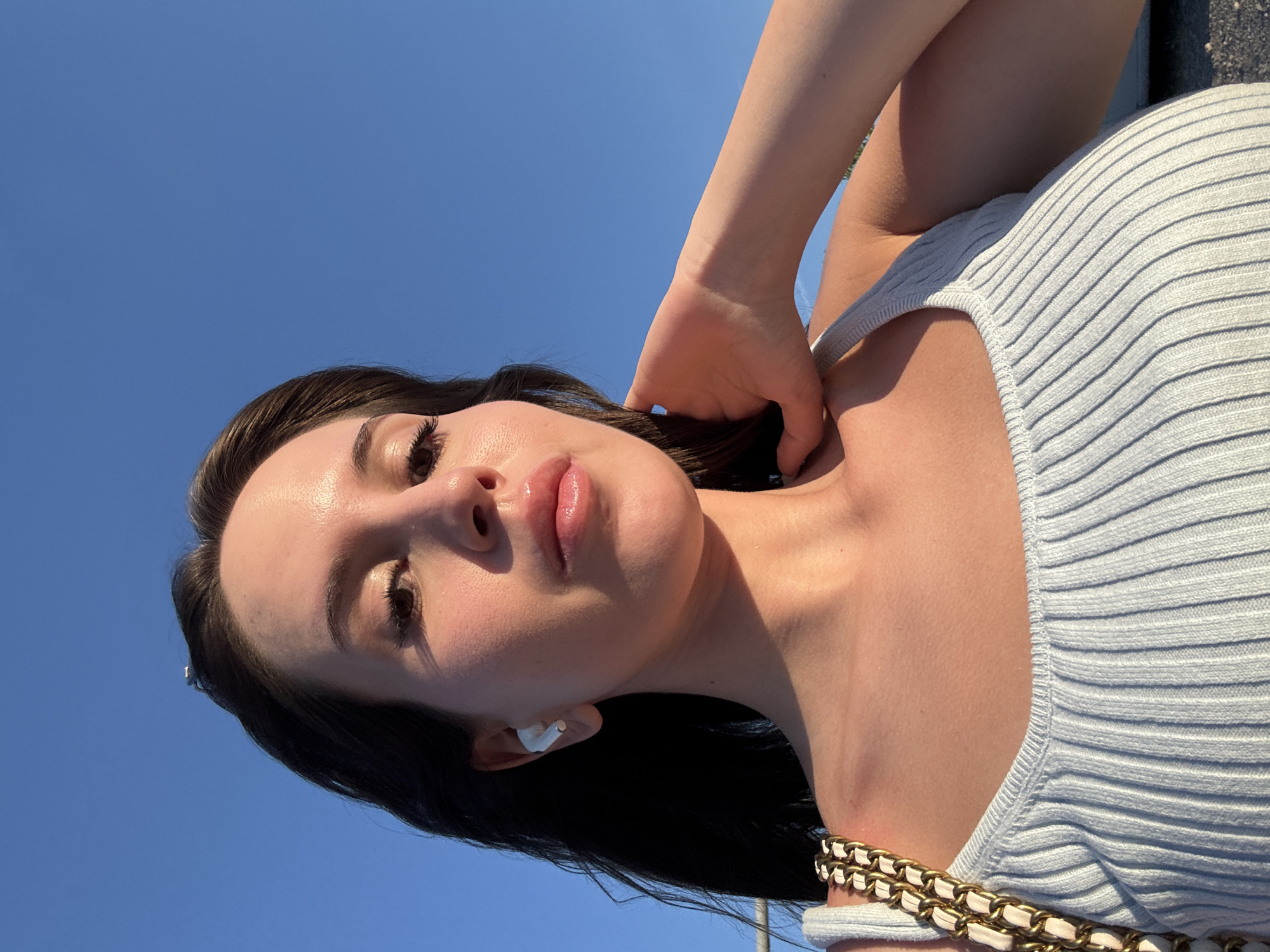 The media file is a close-up, vertically oriented portrait of a young woman. She has dark hair, fair skin with visible makeup on her eyes and lips, and is wearing a light-colored, ribbed top with a gold and white chain strap. Her left arm is raised and her hand is resting on the back of her neck or hair. The background is a clear, bright blue sky, suggesting it is daytime with fair weather. The lighting indicates a sunny day, possibly late afternoon due to the warm glow on her skin. The woman's expression is neutral or slightly posed, with her gaze directed slightly off-camera. A small, white earbud is visible in her left ear. There are no visible text elements in the image. The overall impression is a casual outdoor selfie.
