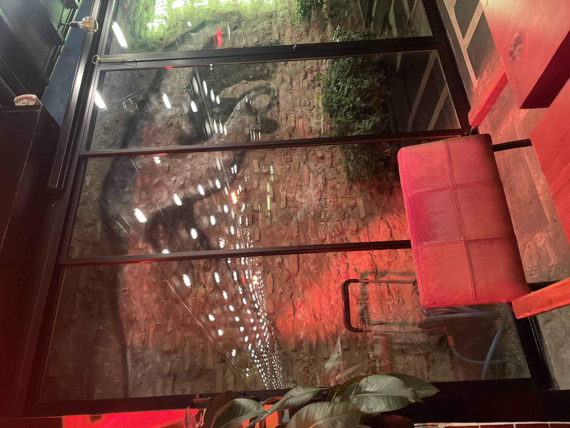 The image shows an interior view through a glass window in what appears to be a commercial or residential space. Outside, there is a brick wall illuminated with a string of lights leading into the distance. There is also some greenery visible at the top of the wall. Inside, a red leather chair is visible, along with a red wooden structure, potentially a table or another piece of furniture. The reflection in the glass window adds depth and complexity to the scene. The exact location in Tbilisi, Georgia, cannot be determined solely from the image without further contextual information.