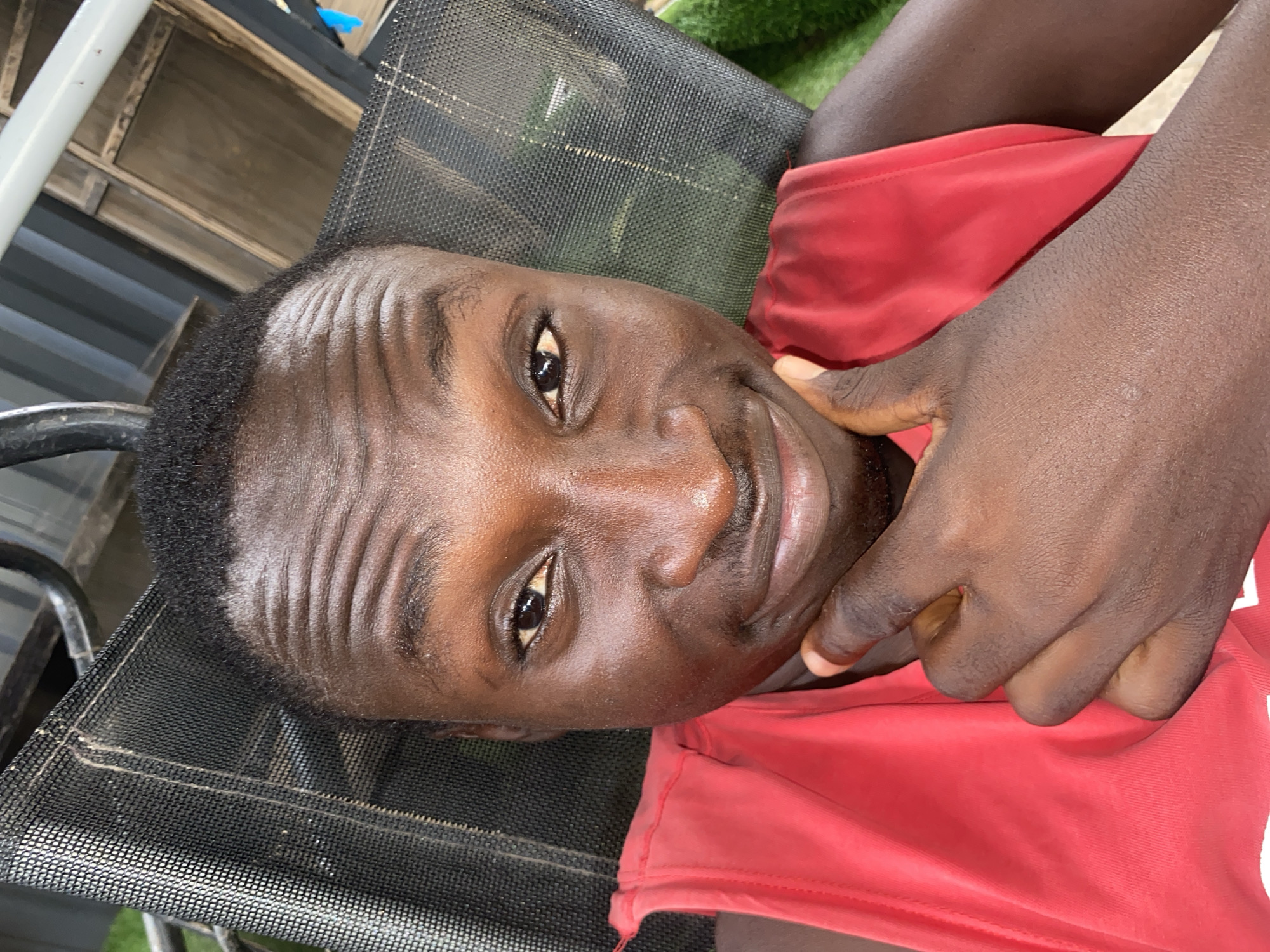 A close-up portrait, oriented vertically, captures a young person with dark skin looking directly at the camera with a soft, friendly smile. They are wearing a red top, likely a t-shirt. The person is positioned as if lying back or reclining on a dark, coarse mesh surface, possibly an outdoor chair.
Their right hand is visible near their face, with fingers gently touching their chin or cheek. Notable features include short, dark hair and several distinct horizontal wrinkles across their forehead. The skin appears somewhat shiny, possibly due to natural oils or bright lighting.
The background elements are somewhat indistinct but suggest an outdoor or semi-outdoor setting. Hints of a white structure are visible on the left, alongside darker, possibly wooden or metallic, structures. Patches of green, which could be grass or artificial turf, are discernible in the upper right and lower left portions of the background. The bright illumination indicates it is daytime. The scene conveys a relaxed and casual moment, potentially in Barden Yana, Nigeria. No text is visible in the image.