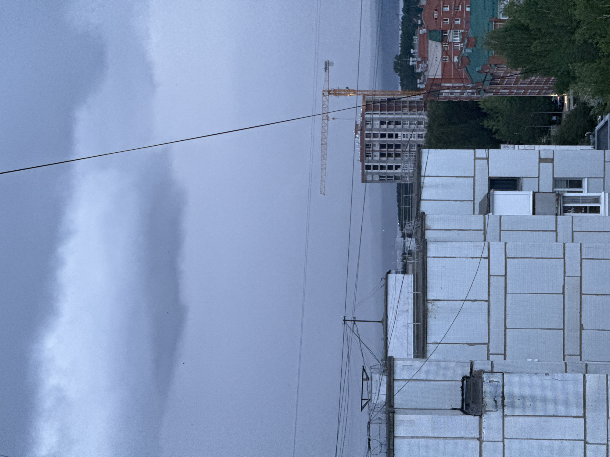 The image presents an urban landscape under a heavily overcast, dull blue-grey sky during late evening or dusk in the city of Chaikovsky, Russia. In the immediate foreground, the upper portions of two light-colored, panel-clad residential buildings are visible, with power lines and communication cables horizontally strung across and diagonally traversing the frame. One building exhibits a damaged section on its left upper facade. In the midground, a multi-story building under construction, primarily concrete or steel framed with multiple open window apertures, stands next to a prominent yellow tower crane. To the right of the construction site, several red brick residential buildings with green sloped roofs are partially visible, interspersed among dark green trees. A narrow street with a vehicle exhibiting illuminated headlights is discernible between the trees. In the distant background, a wide body of water, likely the Votkinsk Reservoir on the Kama River, stretches across the horizon, bordered by a faint, distant landmass. No individuals are depicted in the scene.