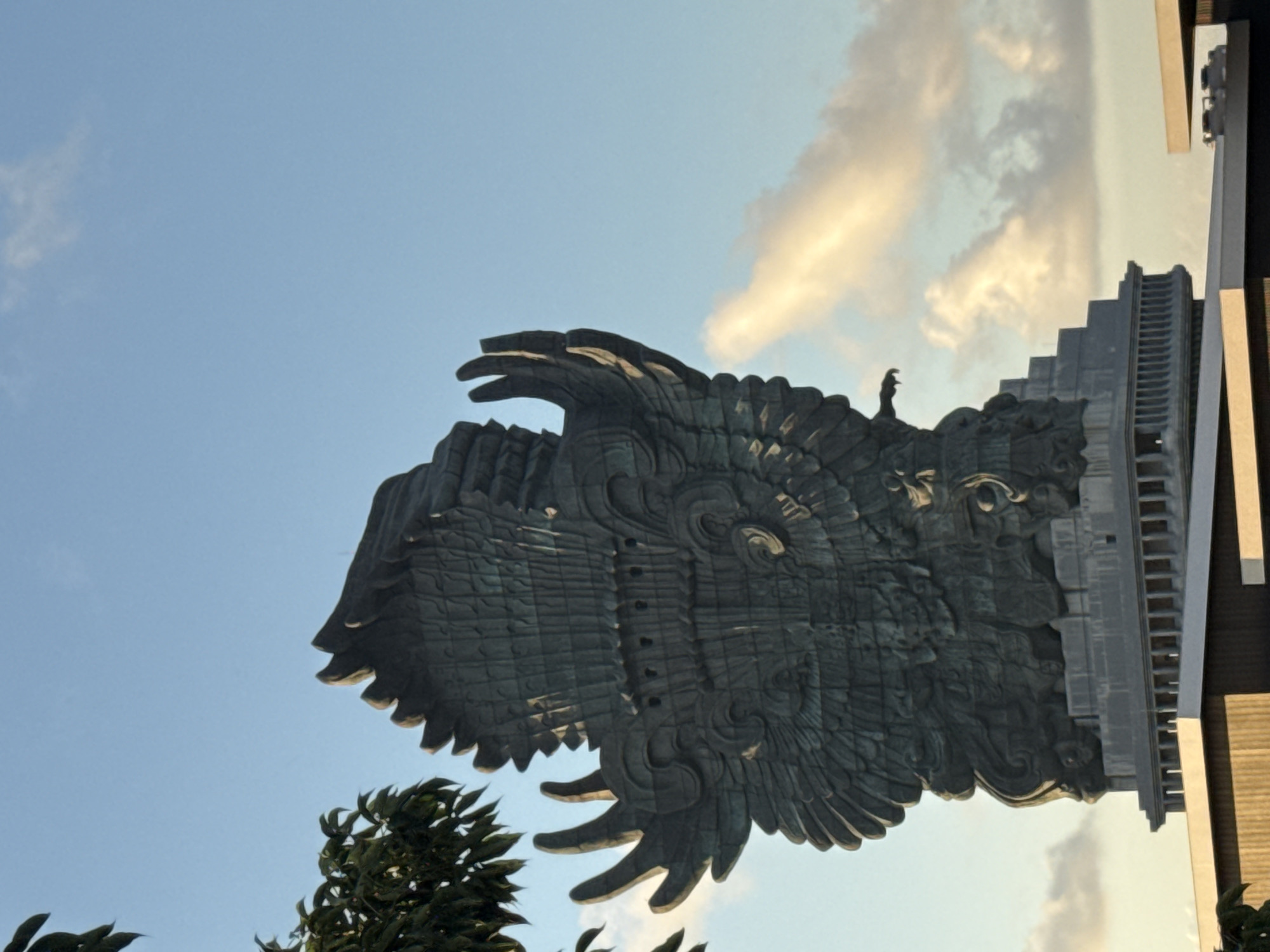 The image captures a low-angle view of the Garuda Wisnu Kencana statue in Ungasan, Indonesia. The statue, massive and ornate, depicts Garuda, the mythical bird. It is situated against a partly cloudy sky and framed by some green foliage in the bottom-left corner. Part of the structural support or a building is visible on the right side. A small figure is perched on the statue's head. The statue is viewed from a skewed perspective.