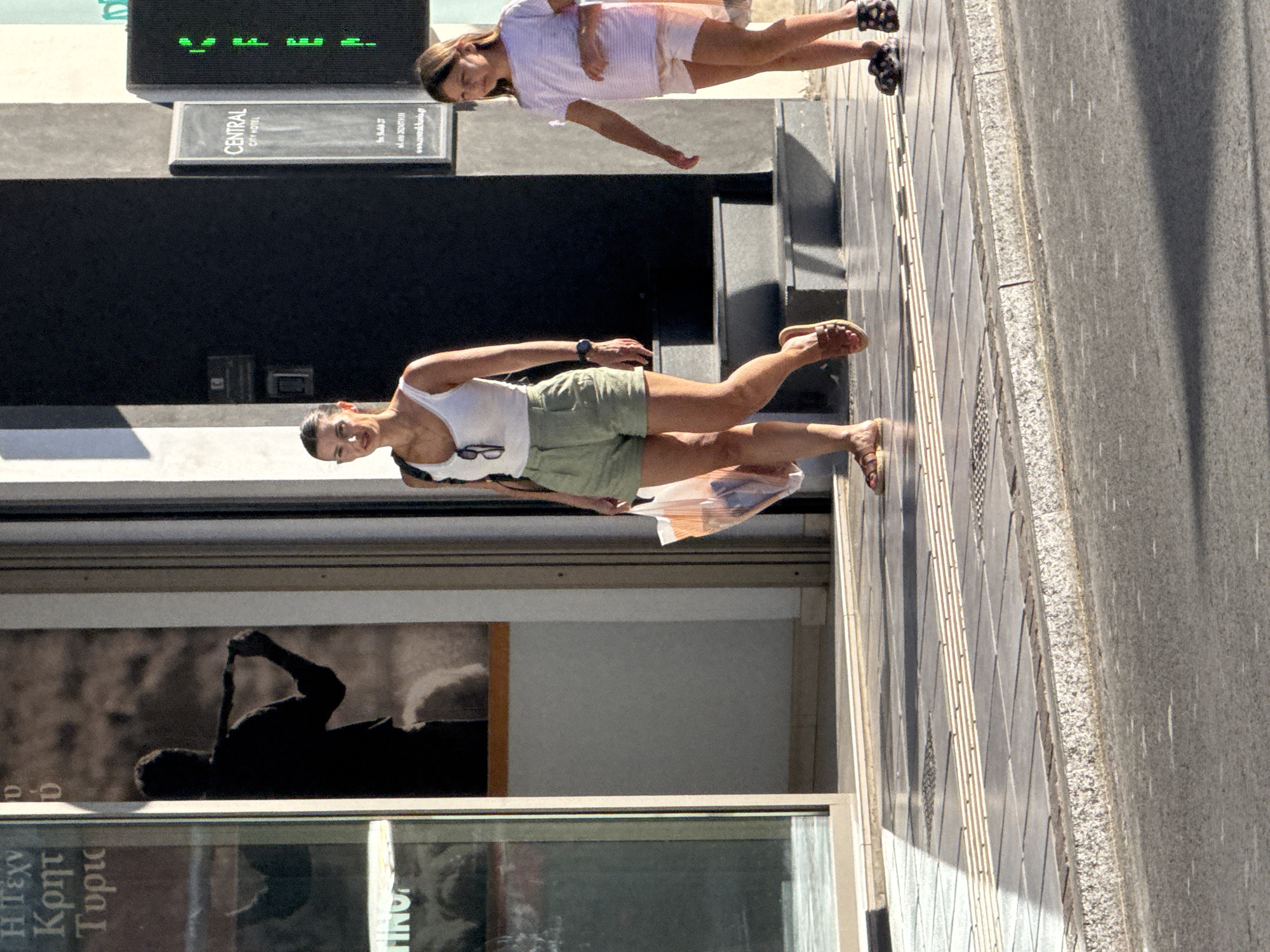 In Chania, Greece, on a sunny day, two females are walking on a sidewalk. The female in the foreground, wearing a white tank top and olive green shorts, is carrying a clear plastic shopping bag. She has dark hair pulled back and is wearing brown sandals. She has a watch on her left wrist. To her right, a younger female in a striped t-shirt and light pink shorts is walking. She has long brown hair and is wearing patterned black sandals. They are walking on a sidewalk made of light gray and darker gray square tiles, with raised tactile paving strips. Behind them is a building with a dark entrance and a sign that reads "CENTRAL CITY HOTEL". A green traffic light is visible on the right side of the frame. The background also shows a large poster or mural on the left, depicting a silhouette of a person holding a stick.