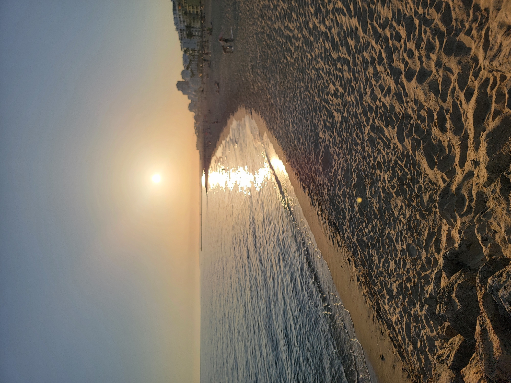 The image depicts a wide sandy beach in Quarteira, Portugal, at sunset. The sun, a bright orb, is low in the sky, casting a warm, golden light that reflects off the calm sea and the wet sand near the shoreline. The water exhibits gentle waves lapping at the beach, leaving a thin line of foam. The sand is textured with numerous footprints and small dunes, indicating recent activity.
Along the coastline, a row of multi-story buildings, likely apartments or hotels, forms a distinct urban backdrop. Palm trees are visible interspersed among the buildings, typical of a coastal resort town. Several individuals are scattered across the beach, engaged in various activities such as walking, sitting, or playing, though they are small in the frame and not the primary focus. The overall scene conveys a serene and tranquil atmosphere characteristic of an evening at the beach. The foreground shows a rocky area at the edge of the beach, providing a natural boundary.