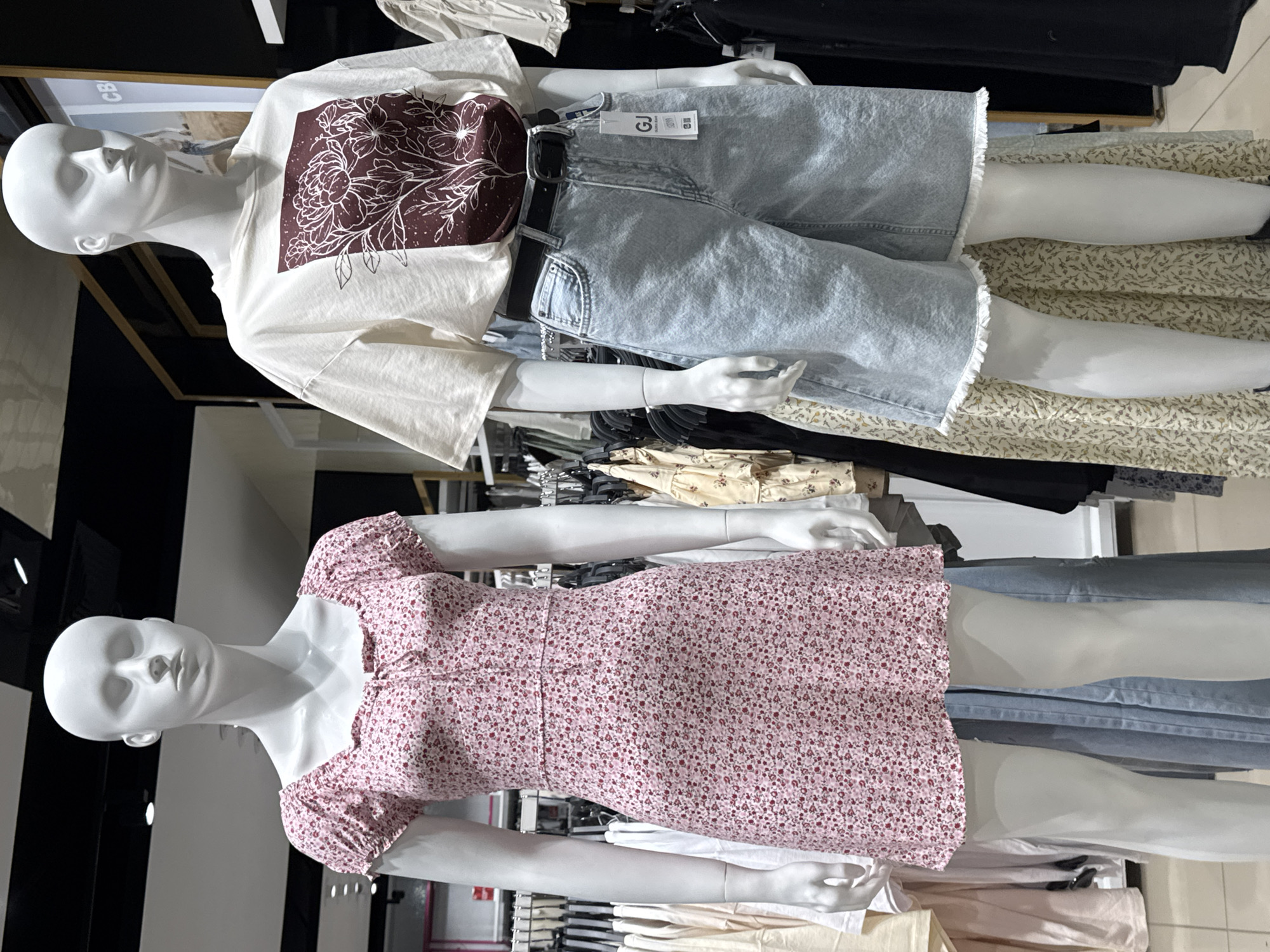 The image shows two mannequins in a clothing store. One mannequin wears a white t-shirt with a floral design, denim shorts, and a black belt. The other mannequin wears a short-sleeved, floral-print dress. The store appears to be in Perm, Russia.