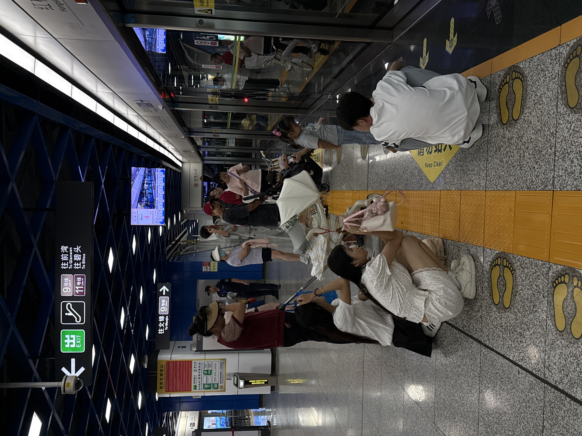 The image depicts a subway platform in the Nanshan District, China. A group of people are waiting for a train, with some seated on the ground and others standing. Two young girls in white outfits are crouched down, facing away from the camera, while a young boy in a white t-shirt is also crouched down, looking towards the train doors. Several individuals are positioned near the platform edge, observing the approaching train. Other passengers, including adults and children, are visible in the background, some with strollers. The platform features yellow tactile paving for the visually impaired and signage indicating train lines and directions. The environment is a modern subway station with a clean, tiled floor and overhead lighting. Reflections are visible on the polished floor and the train doors.