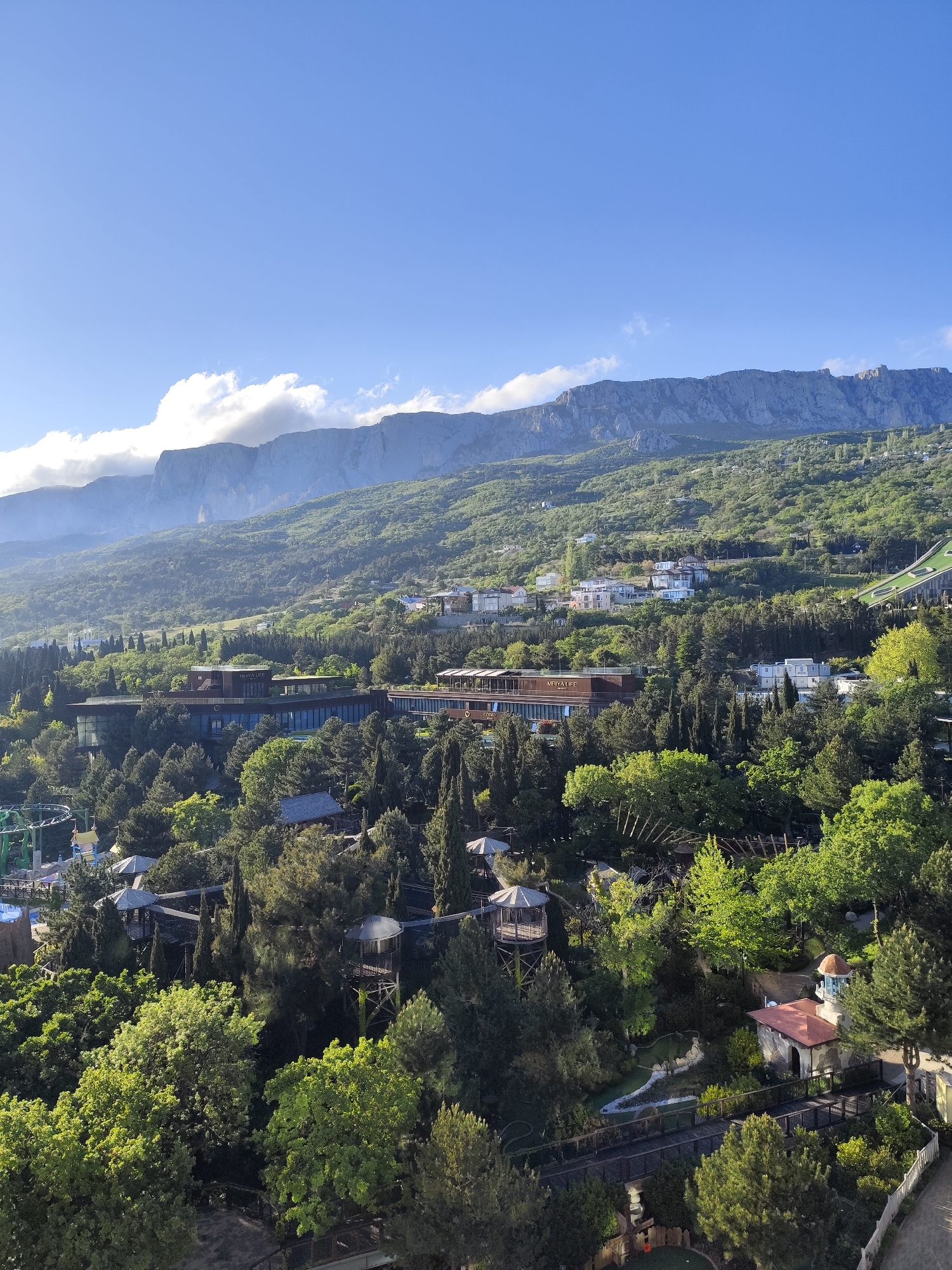 The image depicts a landscape view in the Симеизский поселковый совет, Ukraine, showcasing a forested area interspersed with various buildings and structures.
The upper portion of the image features a prominent mountain range under a clear, blue sky with scattered clouds. The slopes of the mountain are covered in green vegetation, and some areas show rocky terrain.
Below the mountains, the landscape descends into a heavily wooded area populated with a variety of trees and vegetation. Buildings, including a large, multi-story structure, are partially visible among the trees. The structure seems to have a long, horizontal design with visible signage.
In the foreground, there are elevated walkways or bridges, and a mini-golf area with small buildings and a decorative water feature. There is also what appears to be a tower-like structure in the middle ground.