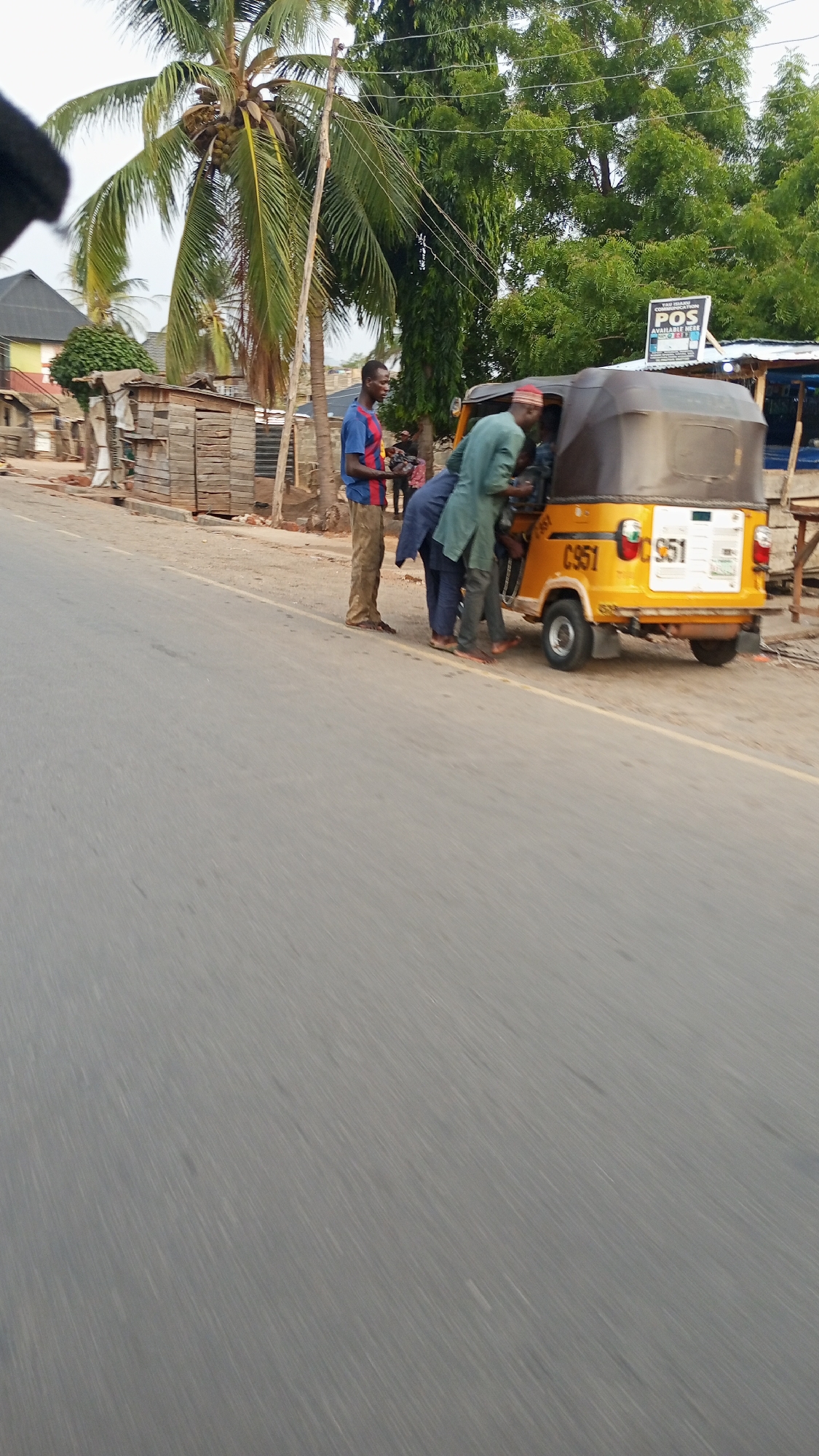 The image captures a roadside scene in what appears to be Zango-ndaji, Nigeria. A yellow and black auto-rickshaw, labeled "C 951", is parked near the edge of a paved road. Three men are near the vehicle: one man in a blue and red shirt and tan pants is standing next to the vehicle, one man in a blue shirt is leaning toward the vehicle, and one man wearing a green tunic and a red cap is positioned at the back of the auto-rickshaw. In the background are some small, rudimentary wooden structures and a coconut palm tree. A sign for "POS AVAILABLE HERE" is partially visible. The foreground is dominated by the asphalt road.