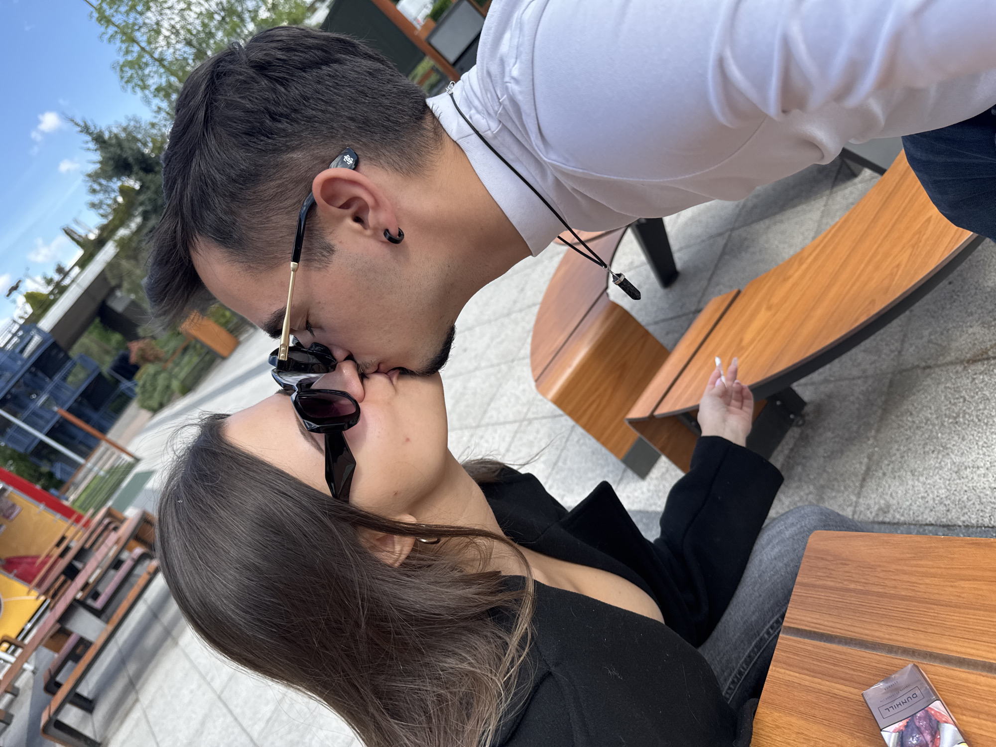 A couple is kissing outdoors, with the man wearing a white shirt and dark sunglasses, and the woman in a black coat and dark sunglasses. The woman is seated at a wooden table and appears to be smoking. A pack of Dunhill cigarettes is visible on the table. The setting appears to be an outdoor seating area in Sofia, Bulgaria, with a building visible in the background.