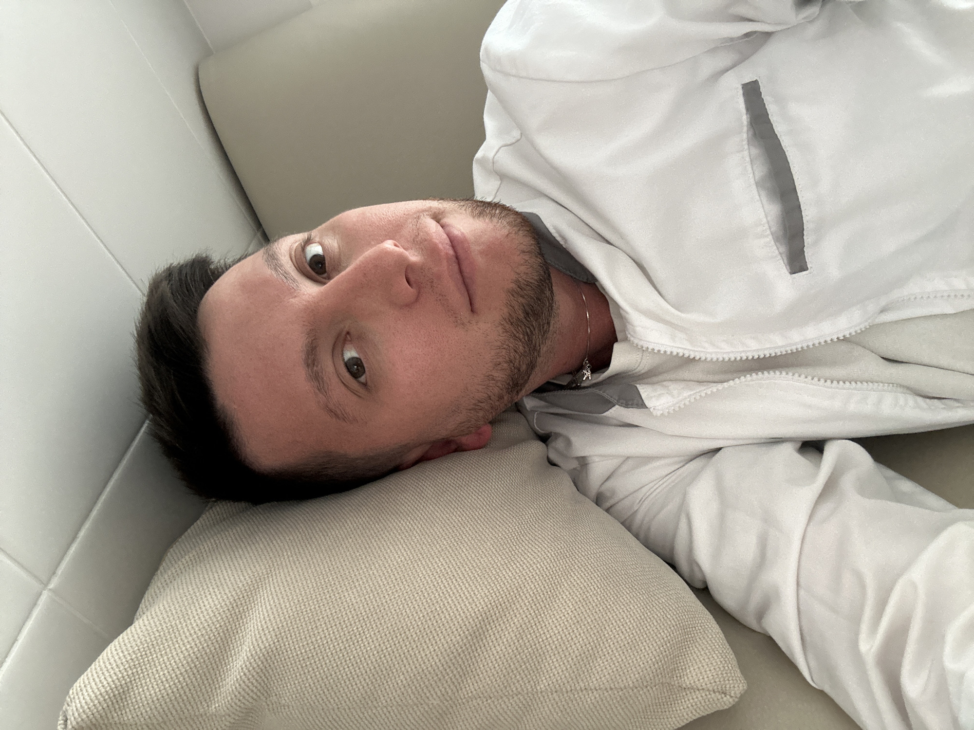 A person is lying on a beige couch. The person has dark hair and a beard and is wearing a white zip-up jacket with a gray pocket. The person is looking directly at the camera with a neutral expression. The setting is indoors, with a white tiled wall visible in the background. There is no indication this image was taken in Moscow, Russia.