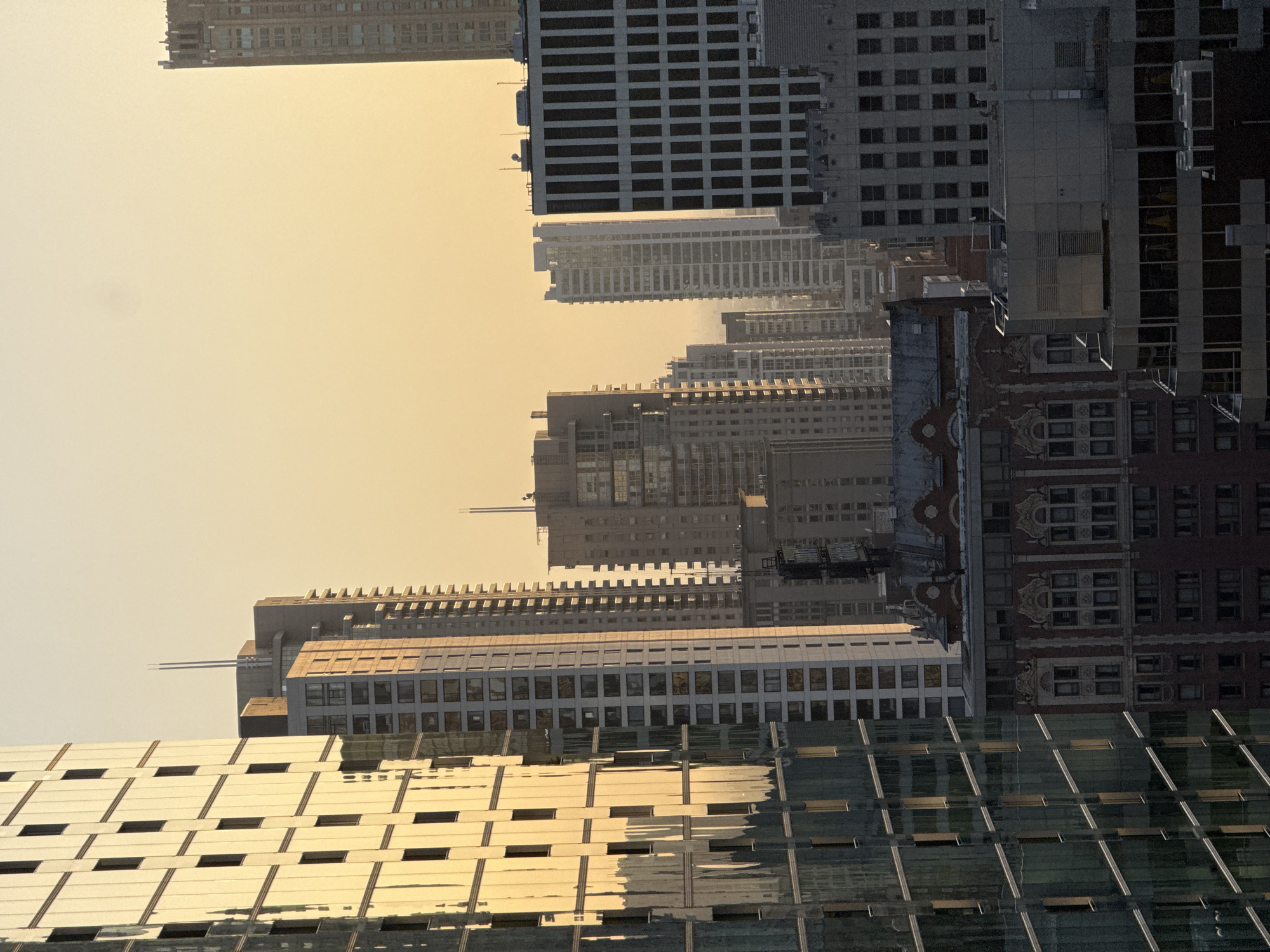 This image captures a skyline view of Chicago, United States of America, likely during either sunrise or sunset, given the warm, hazy glow of the sky. The foreground is dominated by the reflective facade of a modern skyscraper on the left, its glass panels mirroring the golden light and adjacent buildings. To the right of this, a series of tall buildings rise, varying in architectural style and height.
One prominent building in the mid-ground features a dark brick facade with ornate, classical detailing around its windows and roofline. Further into the distance, a dense collection of skyscrapers, many with repetitive grid-like window patterns, recede into the atmospheric haze. The top of one very tall building, possibly part of the older architecture of the city, is visible on the far right, its crown-like structure silhouetted against the sky. The overall impression is one of urban density and the interplay of natural light with man-made structures. There are no people or discernible actions captured in this still image.