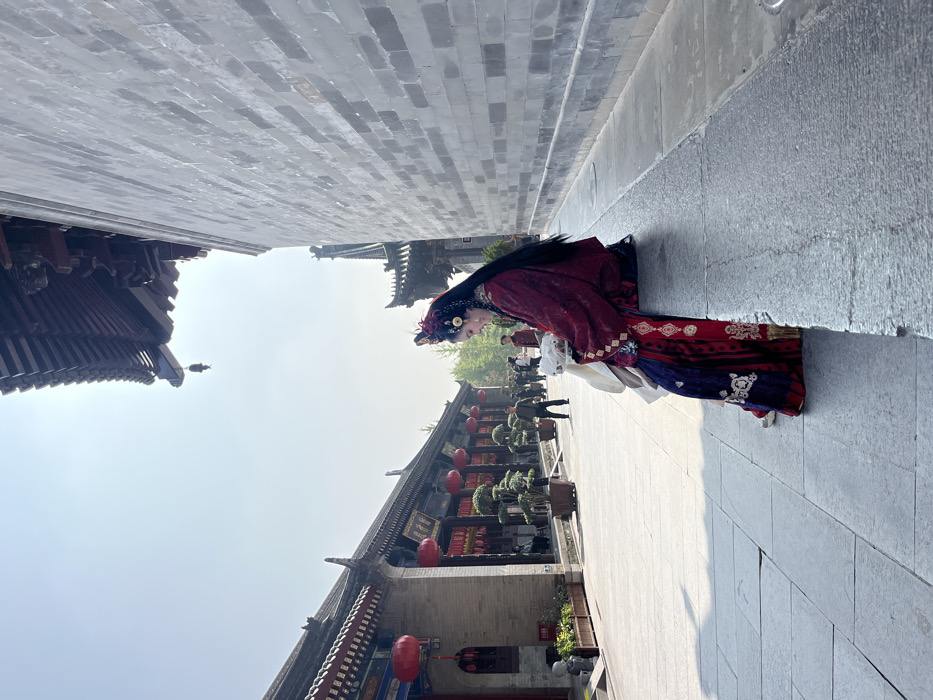 A female individual, adorned in elaborate traditional Chinese attire, is seated on a low stone ledge on the right side of a paved pedestrian street. Her dark red robe features extensive gold and white embroidery, complementing a visible dark blue embroidered undergarment and a white inner layer. A detailed dark headdress with red and gold accents is worn over her long, dark hair. Her hands are positioned in front of her, holding a white object, and she gazes downwards and slightly to the left.
The street, constructed from light-colored stone slabs, extends into the background. To the left, a series of traditional Chinese buildings with dark tiled roofs and exposed wooden structures line the street. These buildings feature red pillars and numerous red lanterns displaying gold Chinese characters. Several potted bonsai-style trees are arranged along this side. A small, light-colored stone statue rests on the ground in the far left foreground. Further down the street, several individuals in casual clothing are visible, walking or standing. The right side of the image is dominated by a tall, textured grey brick wall, capped by a dark tiled roof from which a small bell is suspended. The scene is illuminated by bright daylight, with a clear sky overhead. This location is identified as a cultural pedestrian street, consistent with Datang Everbright City, situated in the Yanta District of Xi'an, China.
