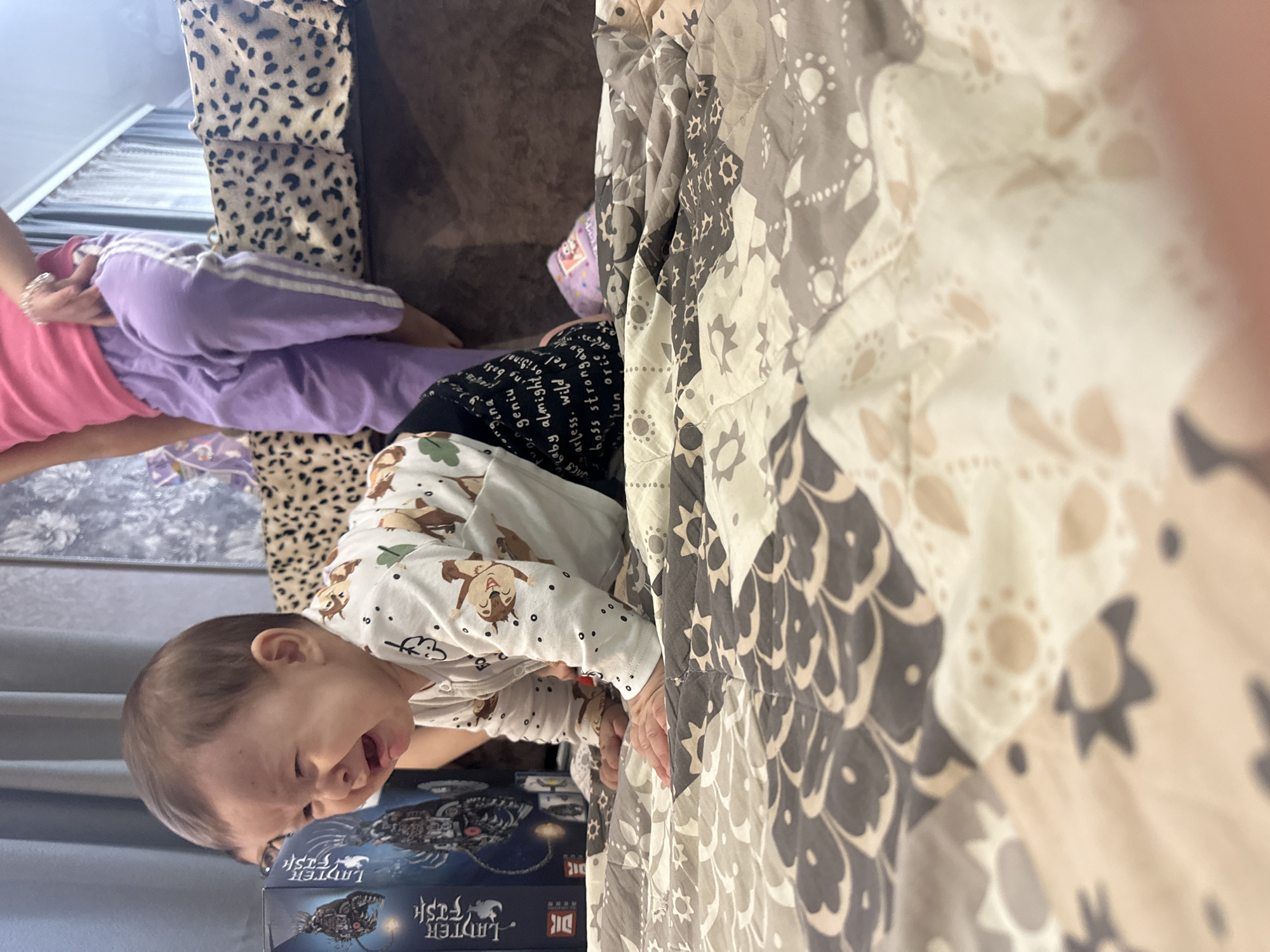 A detailed forensic description of the scene is as follows:
**People:**
* A child is present, wearing a white onesie with an animal pattern, appearing distressed with their mouth open.
* Another person, possibly an adult, is partially visible with a pink shirt and lavender pants.
**Subjects:**
* A boxed item with text "LADTER FISH" is near the child.
* A brown couch and a patterned throw blanket are in the background.
**Objects:**
* Boxed "LADTER FISH" game.
* Brown couch with a decorative animal-print pillow.
* Bed with patterned blanket.
**Actions:**
* Child is crying/making noise.
**Environment:**
* Indoor setting, potentially a living room or bedroom.
**Other:**
* The image is taken at an angle with the camera being rotated to the right.
* The photograph appears somewhat unfocused.