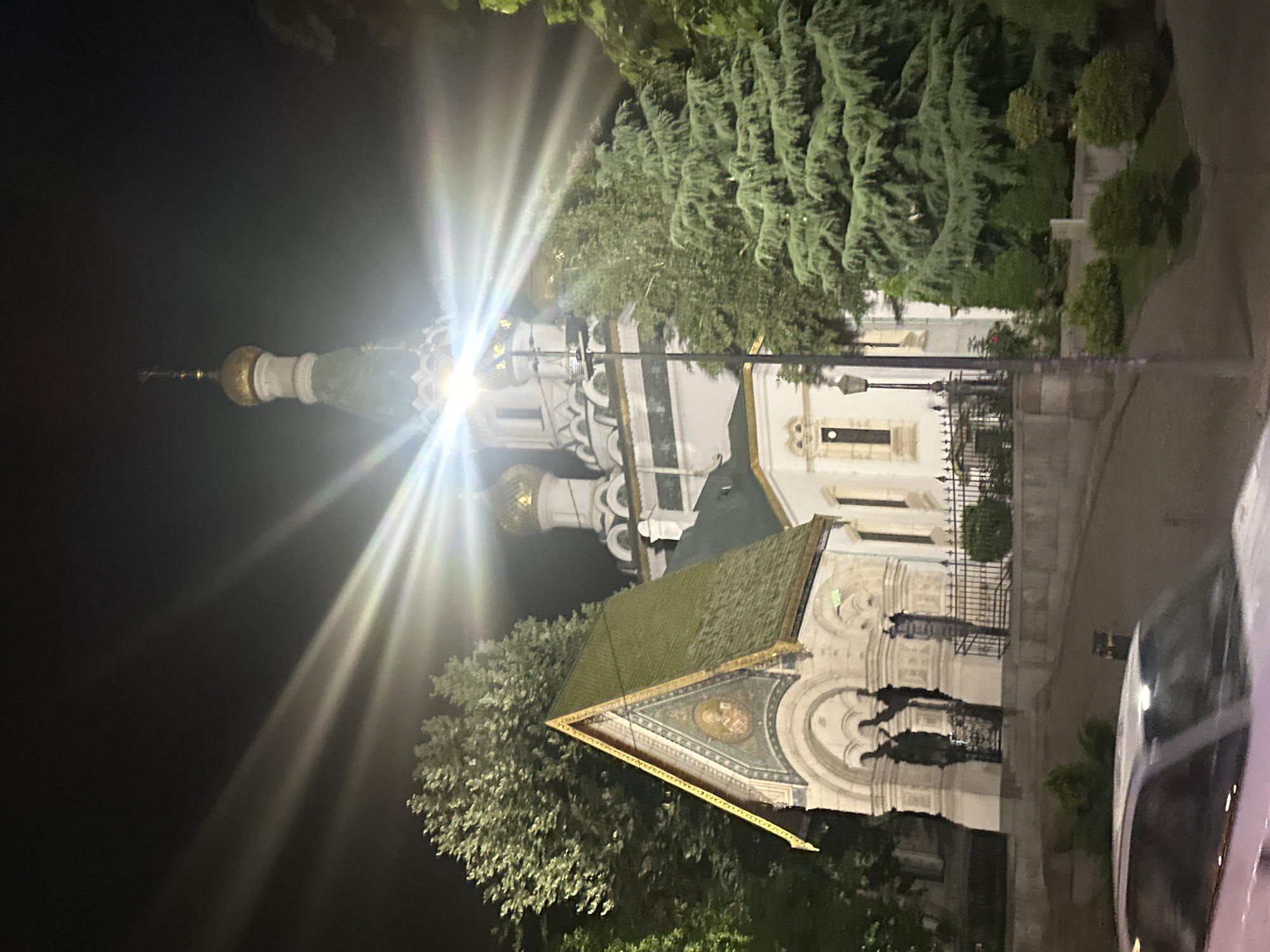 This is a nighttime photograph of a white church with gold domes and a tall steeple, illuminated by bright lights that create lens flares and streaks. To the left of the main church structure is a smaller building with a green, triangular roof adorned with gold trim. In the foreground, the hood and windshield of a white car are visible. The environment includes trees and shrubs, with a dark sky overhead.