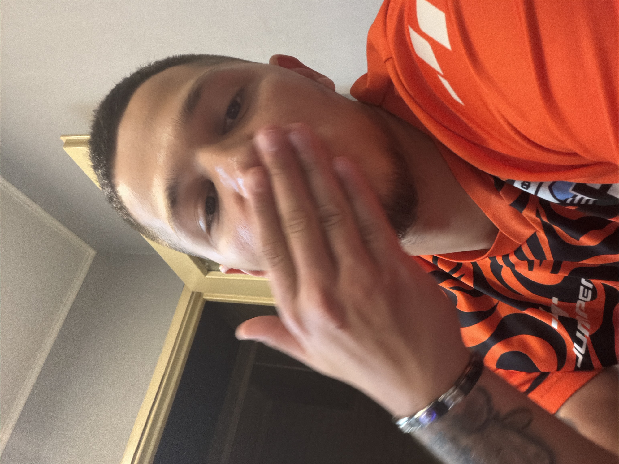 A man, with short dark hair and a light beard, is captured in a close-up selfie. He is wearing an orange t-shirt with black and white tribal-like patterns and a white logo on the chest. His left hand is covering his mouth, and he is wearing a silver bracelet on his left wrist. He has tattoos on his left arm. The background shows a wall and a doorway with a golden frame. The lighting is indoor and appears to be artificial. The man's expression and pose suggest he is blowing a kiss.