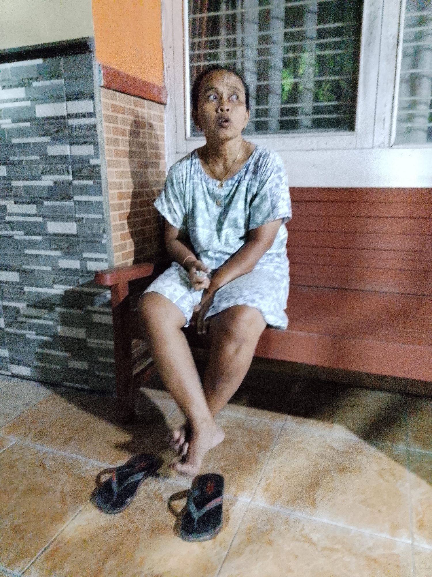 A woman is sitting on a wooden bench outdoors. She is wearing a light-colored, patterned shirt and shorts. Her legs are crossed. Two pairs of flip-flops are on the tiled ground in front of her. She is looking upwards and to her left, with her mouth open as if speaking. The background shows a brick wall and a window with bars. The scene appears to be in a residential area in Kecamatan Pati, Indonesia.
