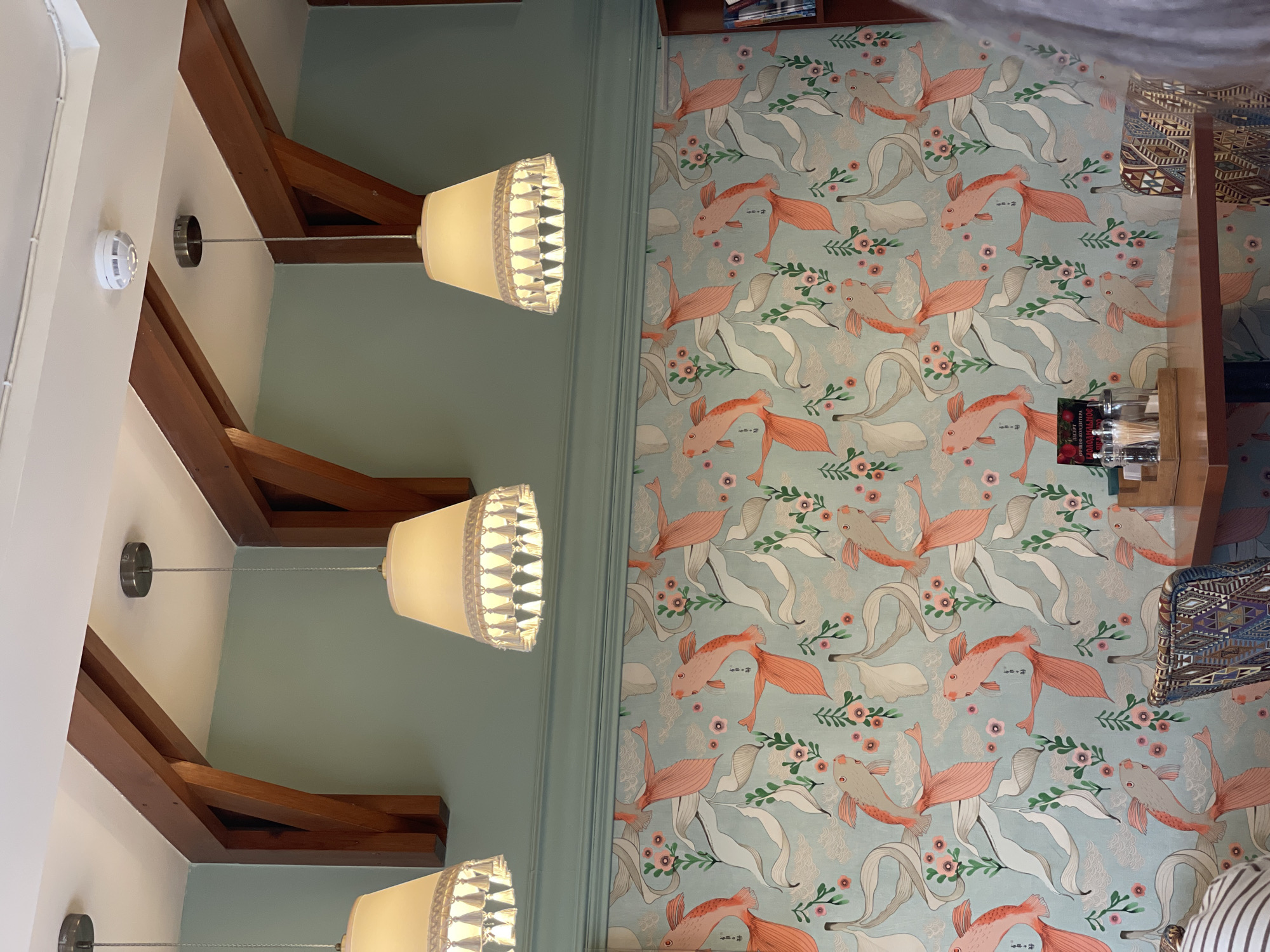 The image shows the interior of a room, likely a cafe or restaurant, with light green walls and a light blue wallpaper featuring a pattern of stylized koi fish and floral elements. Three cream-colored pendant lights with tassel trim hang from the ceiling. Wooden beams support the ceiling. A smoke detector is visible on the ceiling. A wooden table is set with condiments, and two chairs with patterned upholstery are visible. The scene appears to be located in Izhevsk, Russia, based on the provided context.