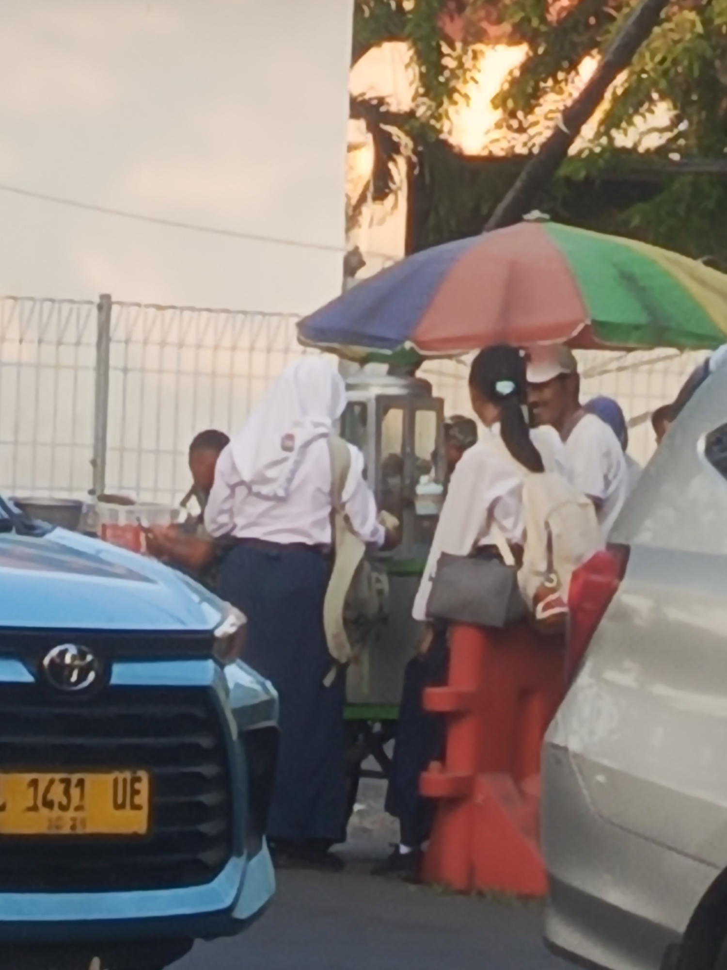 The image depicts a street scene, likely in Surabaya, Indonesia, during the late afternoon or early evening, judging by the warm, soft lighting. A blue Toyota Avanza is partially visible in the foreground, its license plate reading "L 1431 UE". Behind the car, a colorful umbrella, with segments of red, green, and blue, shades a small food stall. Several people are gathered around the stall. On the right, a man wearing a white cap and white t-shirt appears to be talking to someone. To his left, a young woman in a white uniform, possibly a school uniform, with a hijab and a backpack, is interacting with the stall. Another individual, also in a white shirt, stands further left, partially obscured. The background shows a chain-link fence and trees with sunlight filtering through the leaves, suggesting an outdoor, urban environment. Red plastic traffic barriers are also present near the food stall.