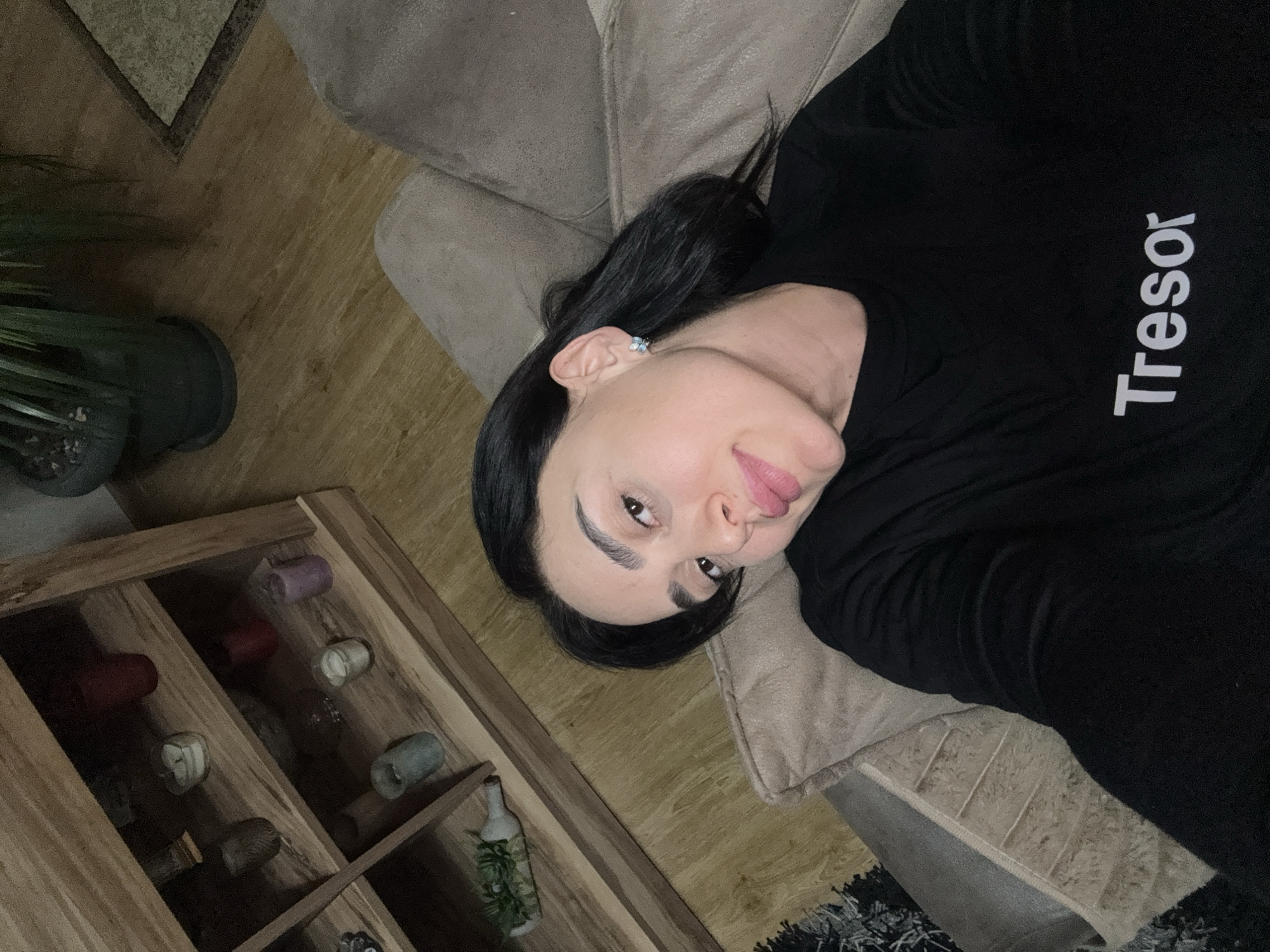 A person is lying on a light brown upholstered couch. The person has dark hair and is wearing a black shirt with white lettering, part of which reads "Treso". The person is facing the camera with their head tilted. In the background, there is a wooden display cabinet with various objects inside, including what appear to be candles and a bottle with greenery. There is also a potted plant to the left of the cabinet and part of a rug visible in the lower right corner. The image appears to be taken indoors, but the location in Tbilisi, Georgia cannot be determined from the provided image.