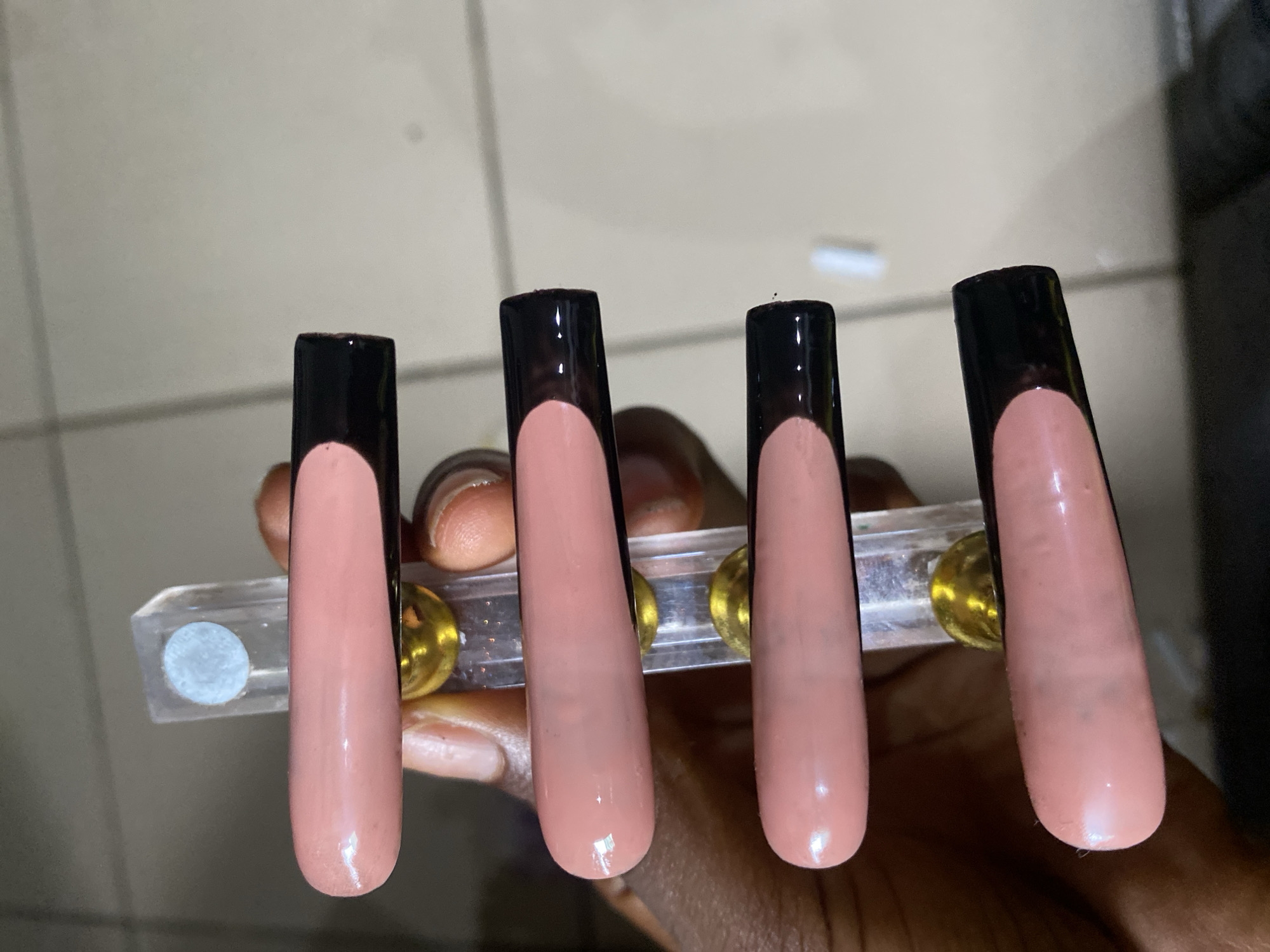 This is a close-up, top-down, vertical shot of a person's hand holding a clear acrylic stand with gold hardware, which displays five long, coffin-shaped artificial nails. The nails are a light pink color with a black French tip. The image is focused on the nails and the stand, with a blurry background that appears to be an indoor setting, possibly a tiled surface. The person's dark-skinned fingers are visible holding the stand, and their nails are also visible at the bottom of the frame. There is no discernible information about the city of Kubwa, Nigeria, in the image itself.