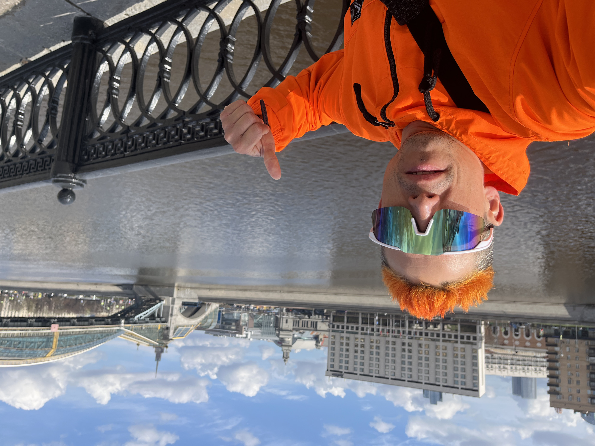 A man with bright orange hair and iridescent sunglasses gives a thumbs-down gesture in front of a river in Moscow, Russia. He wears an orange jacket with black accents. Behind him is a decorative black metal railing. In the background are buildings, a river, and a blue sky with white clouds, reflecting in the water. Visible landmarks include the Moscow International House of Music and the Patriarshy Bridge.