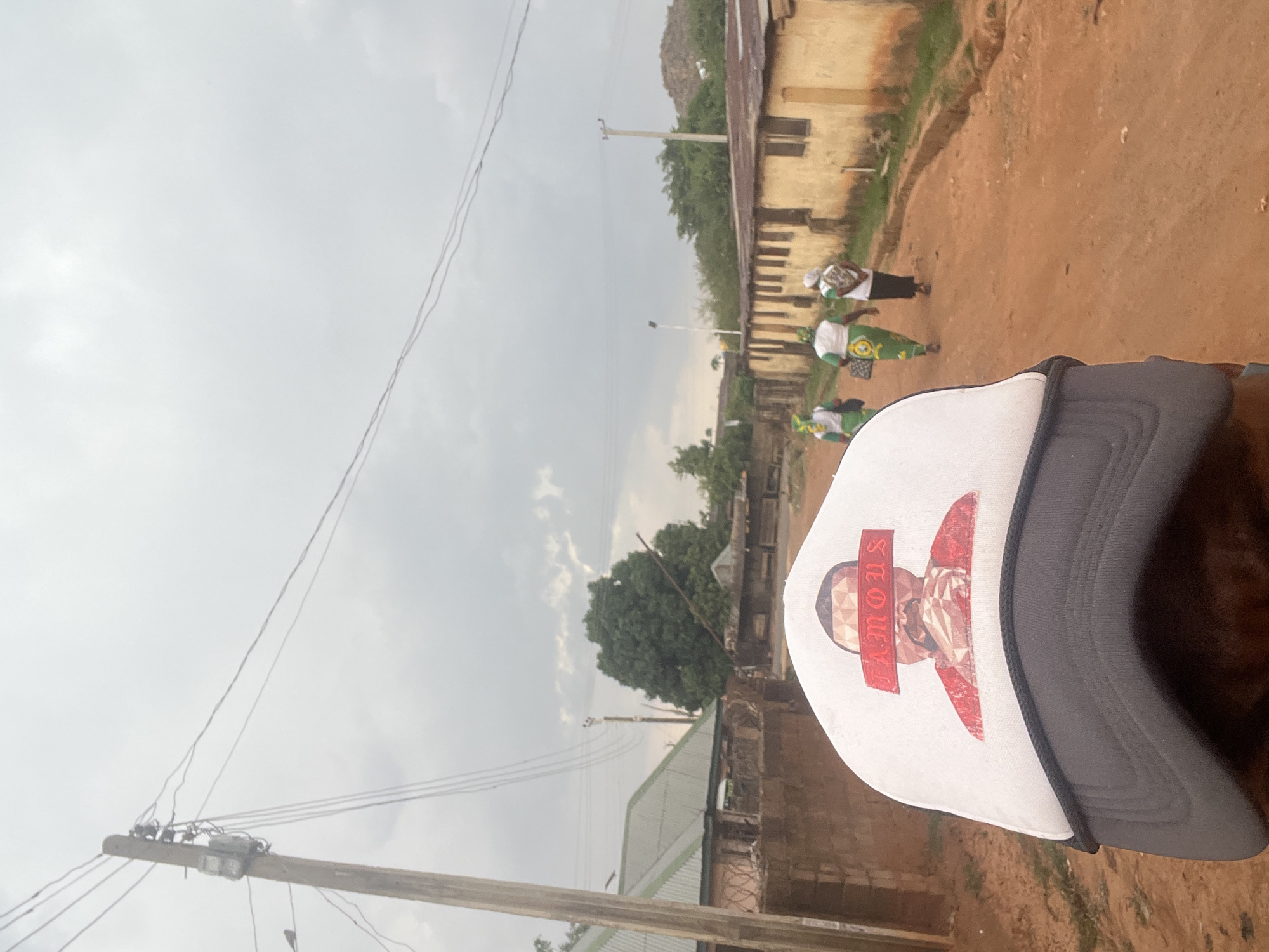 The image shows a person wearing a white and gray hat in the foreground. The hat has a graphic print featuring the word "FAMOUS" across it. Behind the person and hat, there is a dirt road with several people walking on it, some carrying flags. The background includes buildings and trees, with a cloudy sky overhead. Utility poles with electrical wires are visible to the left. The location is Gura Top, Nigeria.