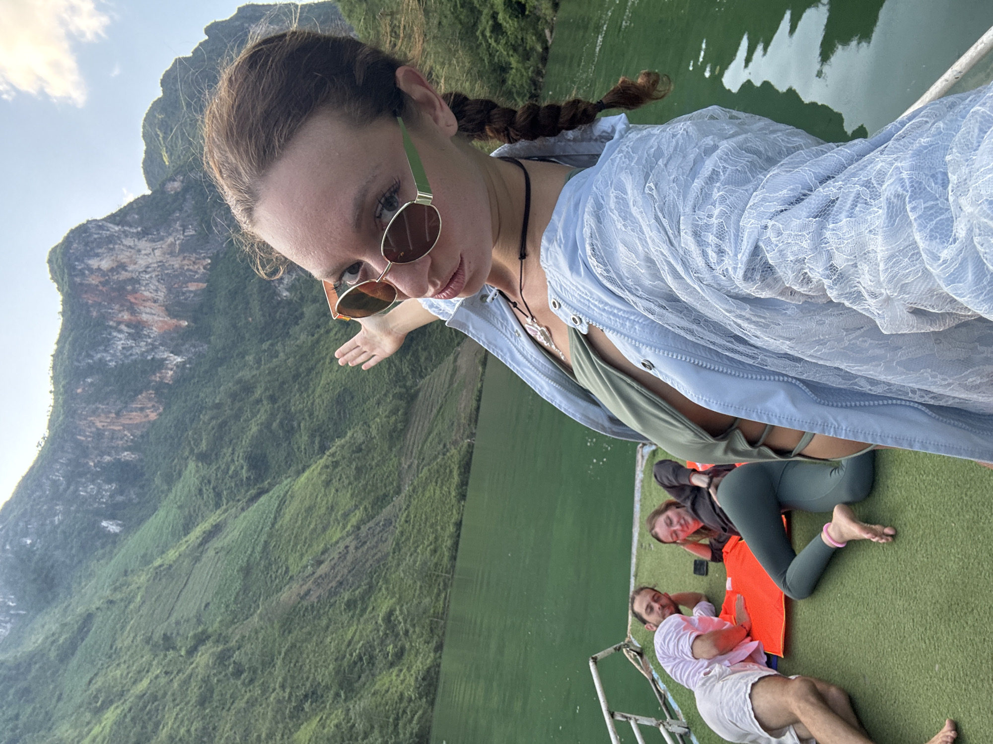 A selfie-style photograph features a woman in sunglasses and a light blue shirt, with a landscape including mountains and a body of water visible in the background. The location is possibly within the Đồng Văn Commune, Vietnam, suggested by the rugged terrain and riverine environment. Three other people, two women and one man, are reclining on what appears to be the deck of a boat. One woman is wearing grey leggings and an orange life vest. The man is wearing a white shirt and shorts. The overall scene appears to be taken on a boat tour in a mountainous, rural area.